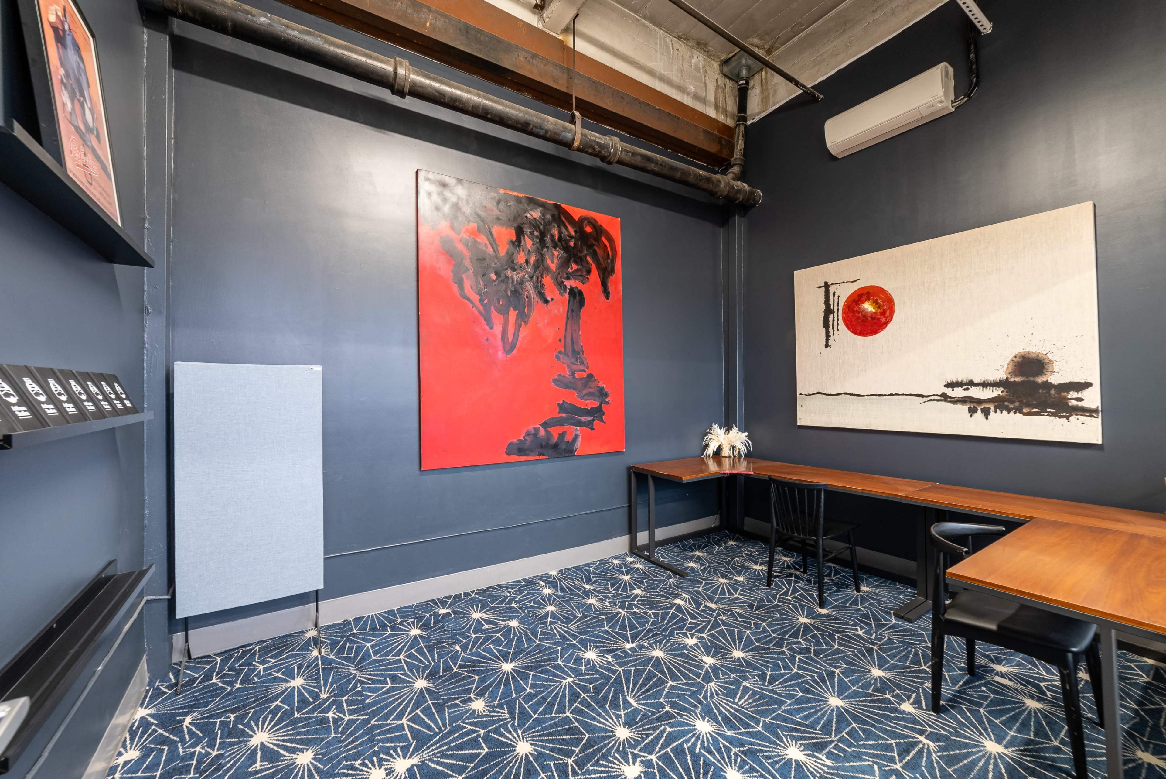A modern interior room with dark blue walls, a patterned carpet, two large abstract artworks, and minimalistic black furniture arranged around a wooden table.