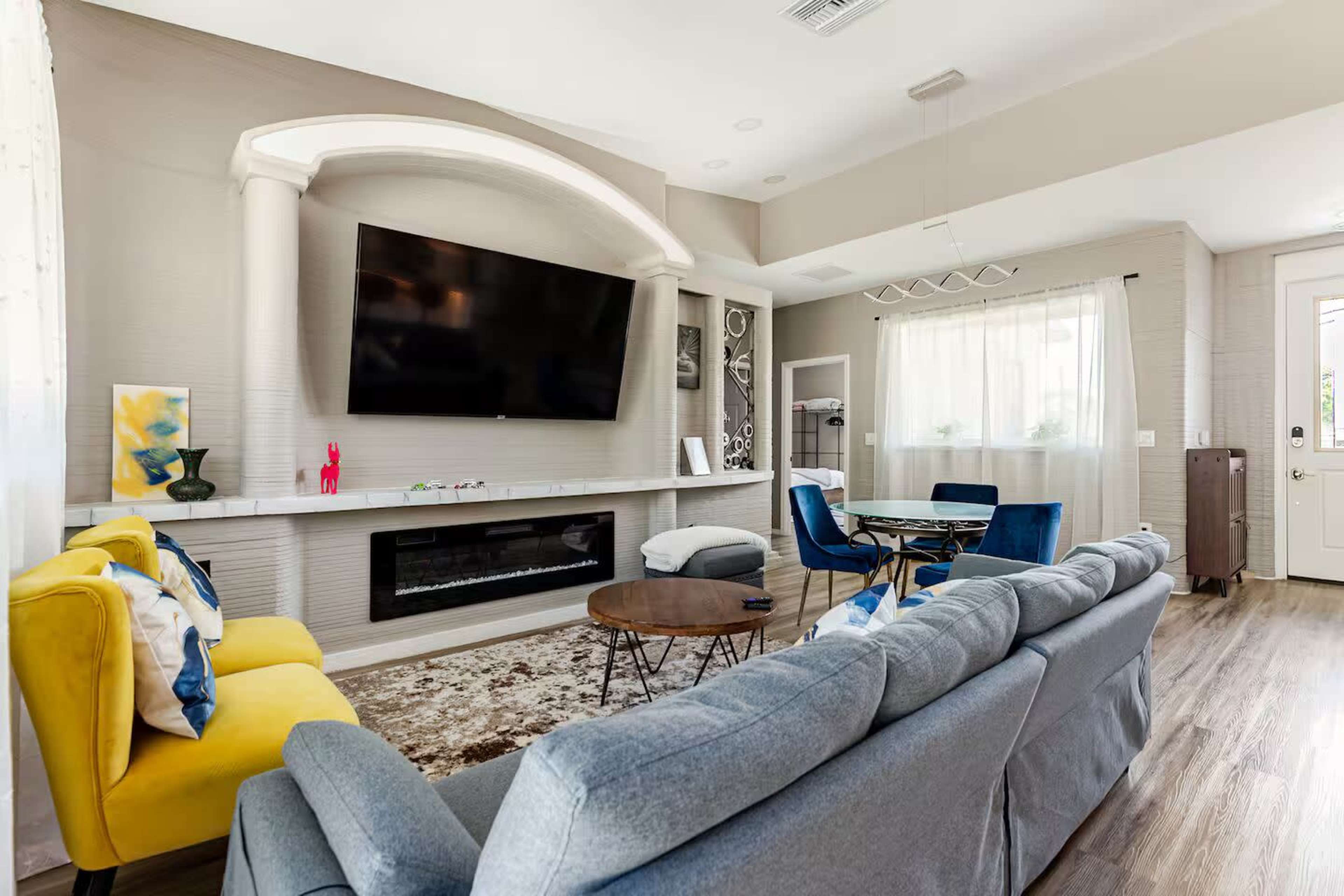 The image shows a modern living room with a gray sofa, a yellow armchair, a large flat-screen TV mounted on the wall, and a dining area with blue chairs and a circular table.