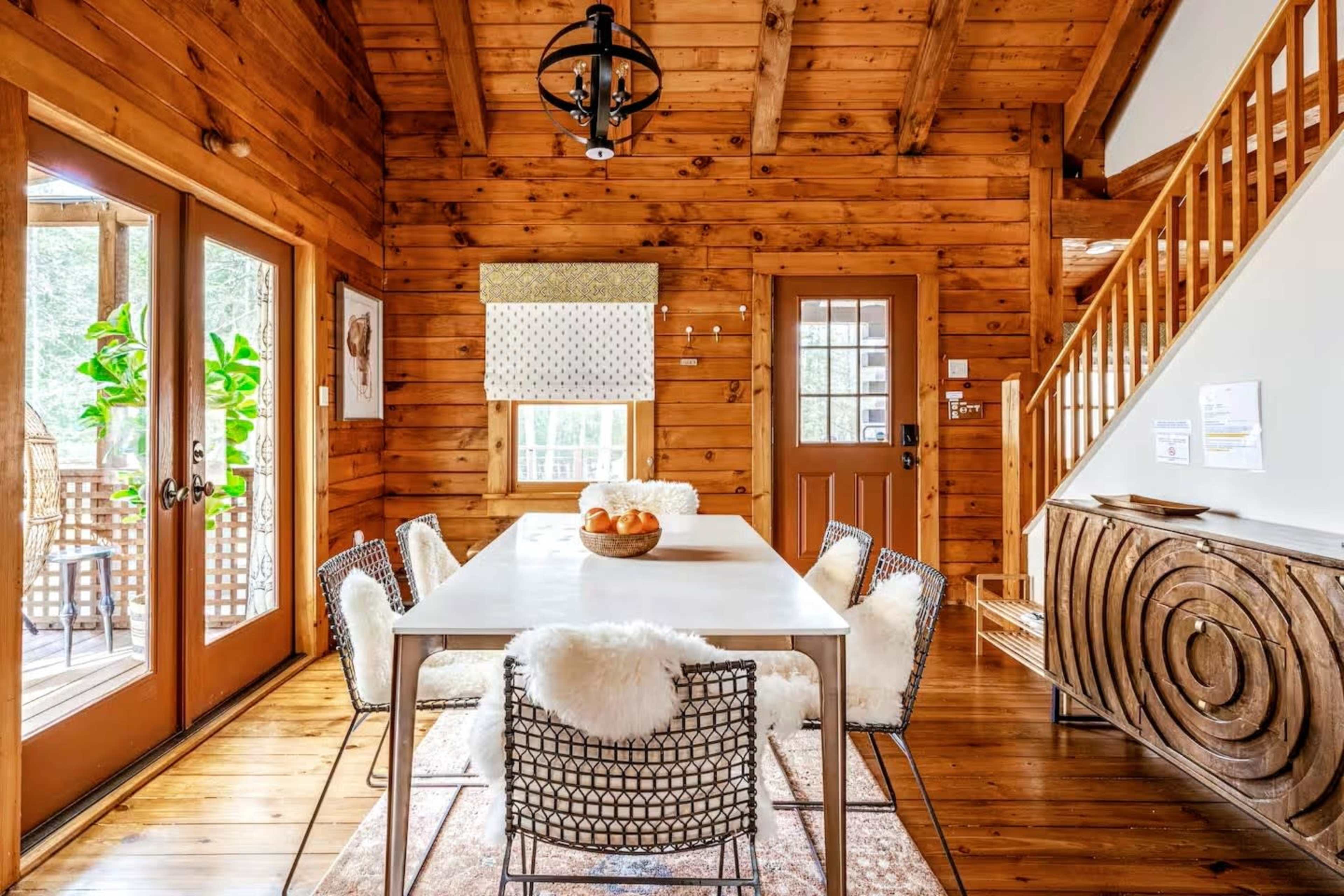 The image shows a wooden interior dining area featuring a white table surrounded by four chairs, with two sets of doors leading to an outdoor space.