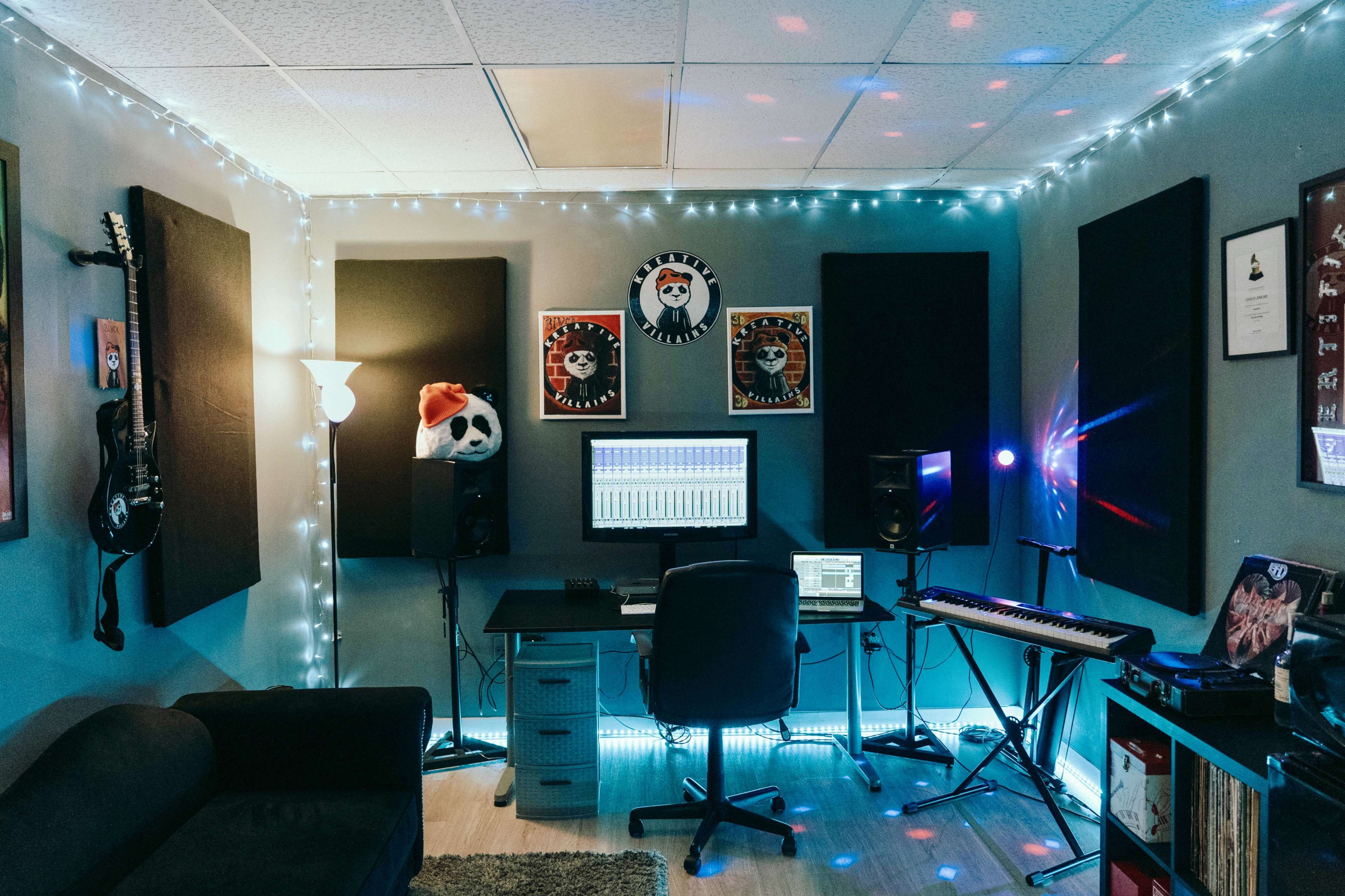The image shows a music studio with a desk set up for production, featuring a computer, speakers, and a keyboard, illuminated by string lights and surrounded by soundproofing panels.