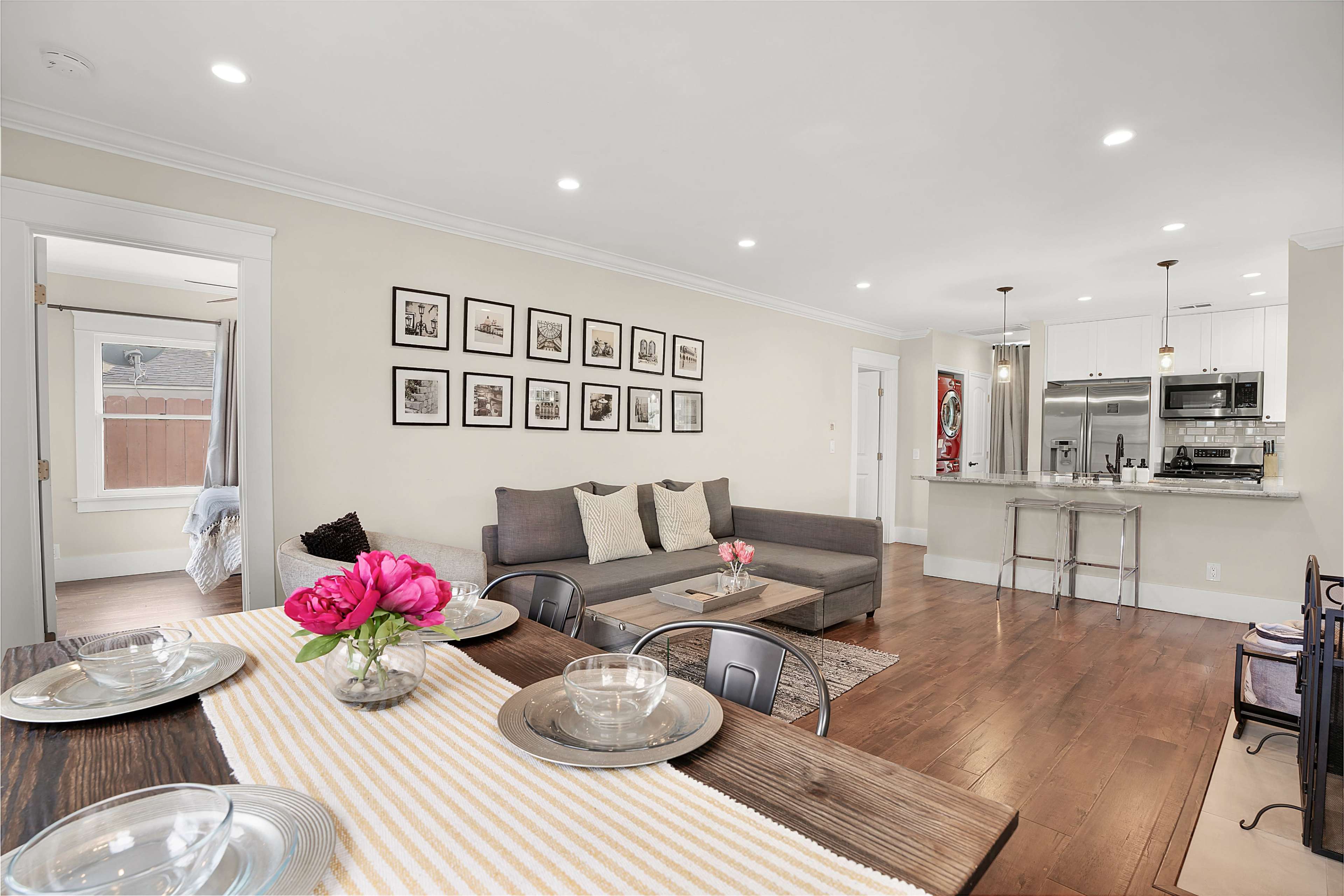 A modern living space features a gray sofa against a wall of framed artwork, with a dining table set for meals and an open kitchen in the background.