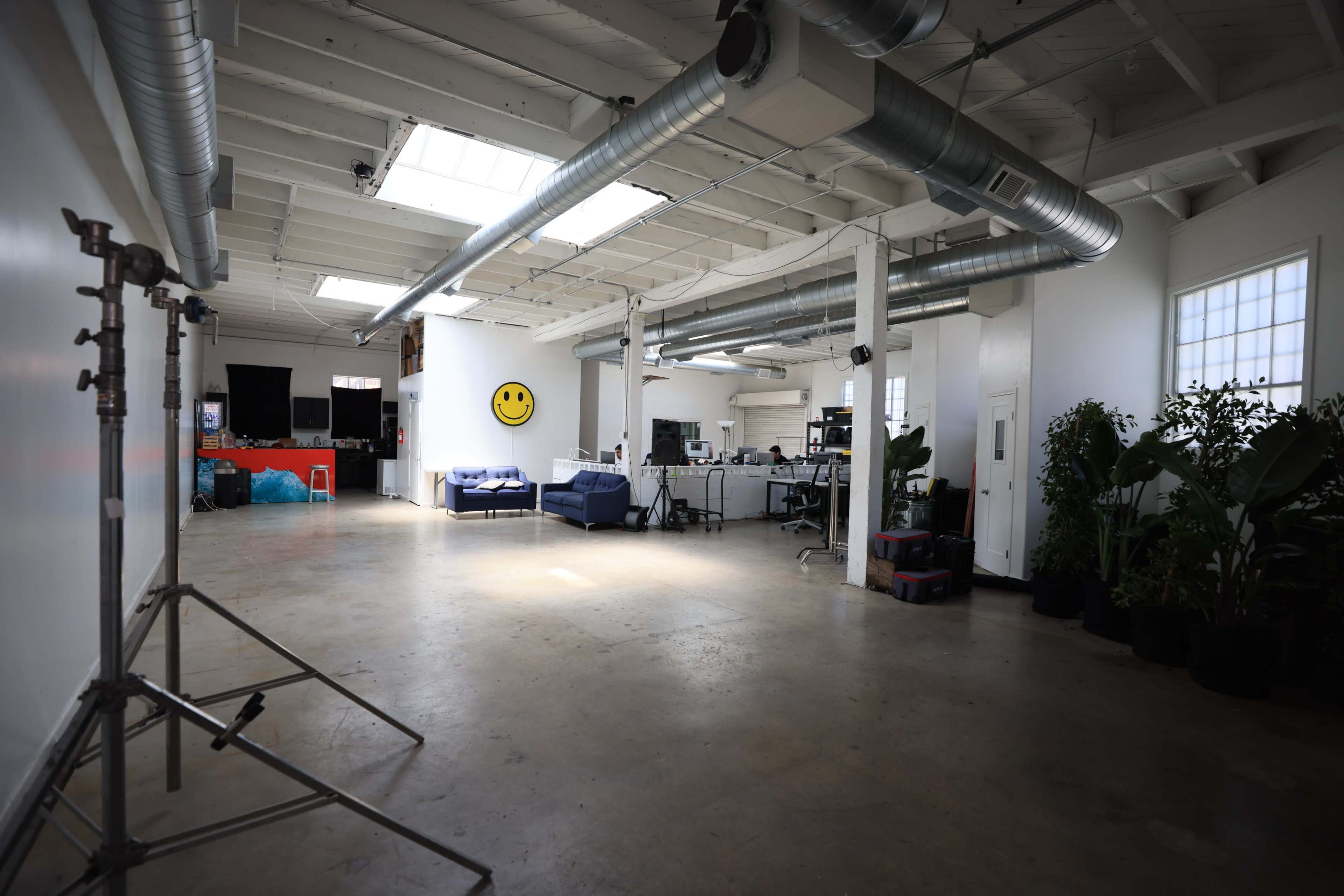 A spacious, well-lit office with large windows, exposed ductwork, and various plants, featuring a blue couch and workstations in the background.