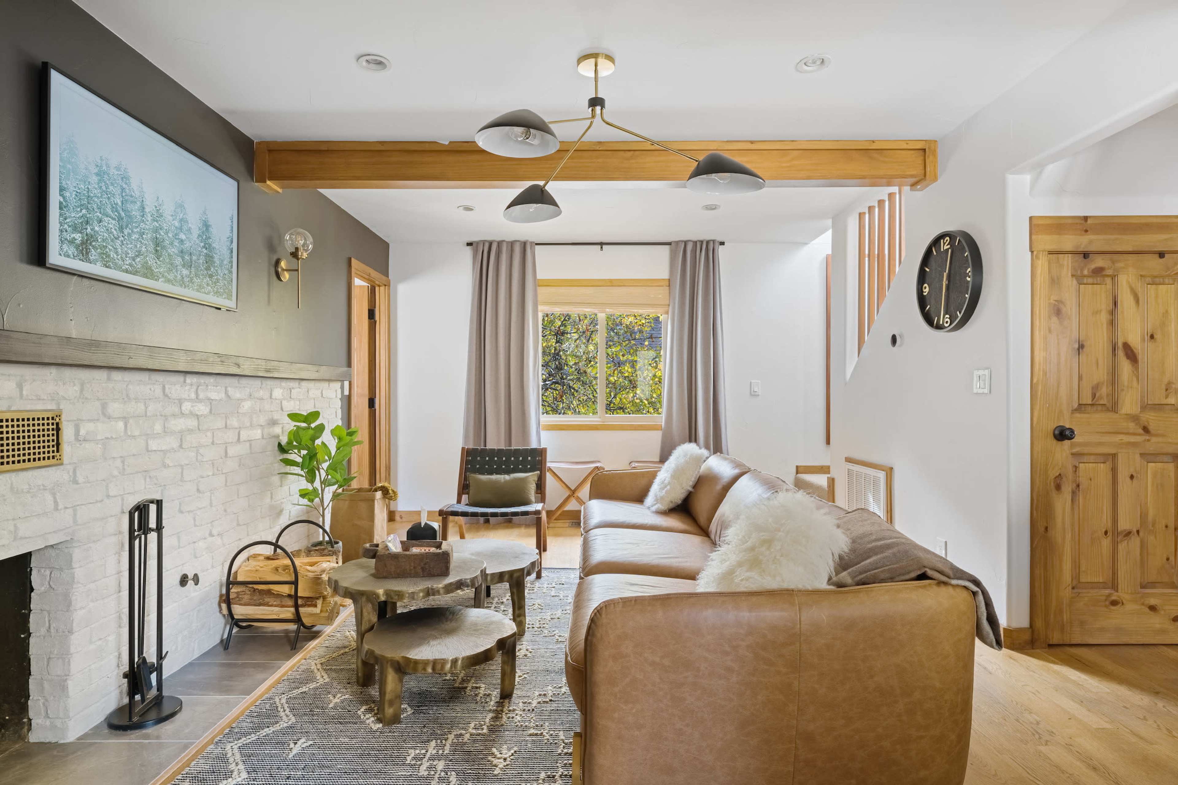 A modern living room featuring a leather sofa, a wooden coffee table, large windows, and a brick fireplace with a decorative wall clock and light fixtures.