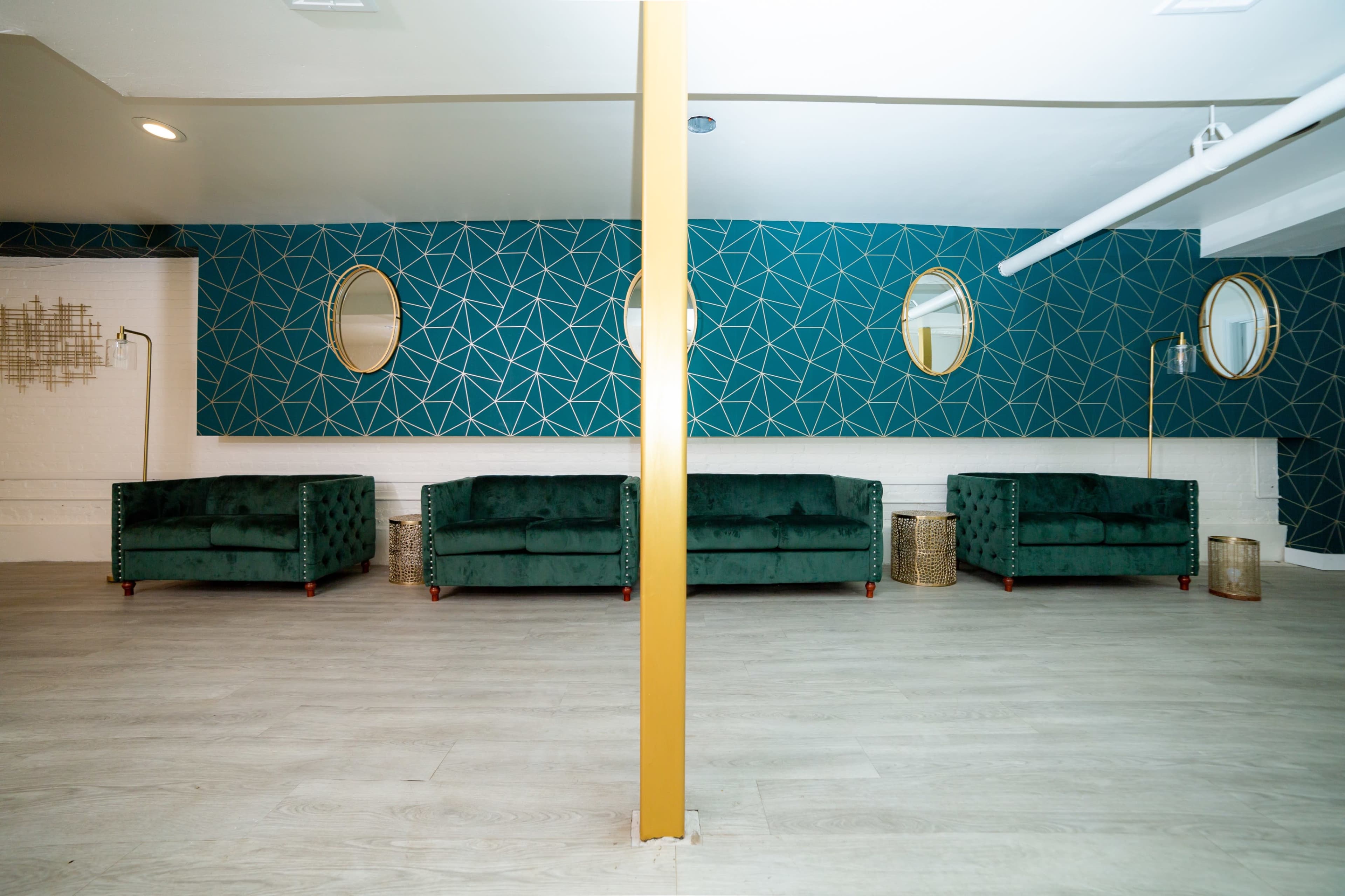 A room with a geometric-patterned teal wall, featuring three green sofas flanked by decorative tables and circular mirrors.