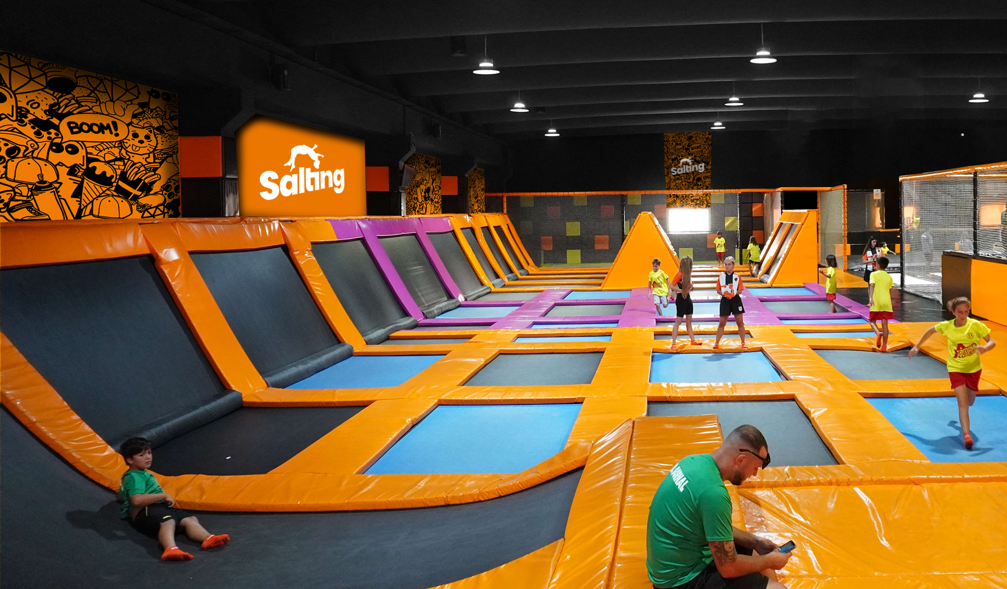 A vibrant indoor trampoline park with orange and black trampolines, colorful wall designs, and several children and adults jumping and exploring the area.