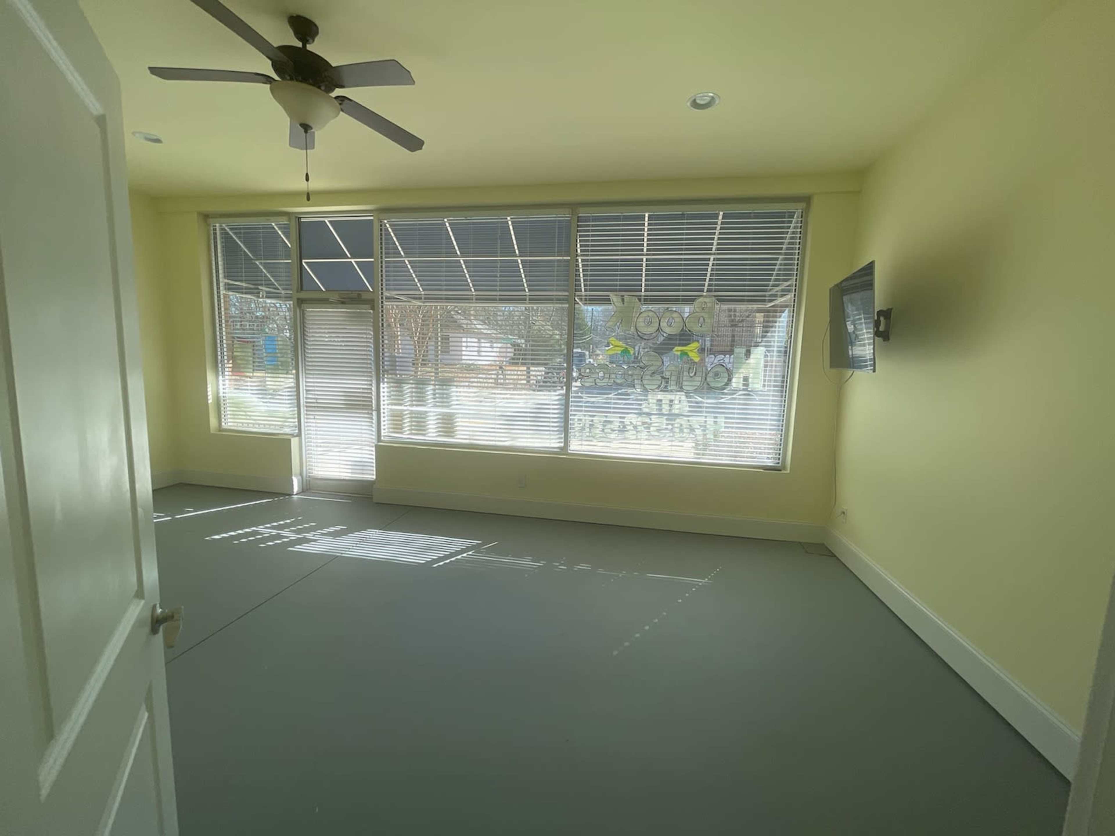 The room features a ceiling fan, large windows with blinds, and a wall-mounted television on a light yellow wall.