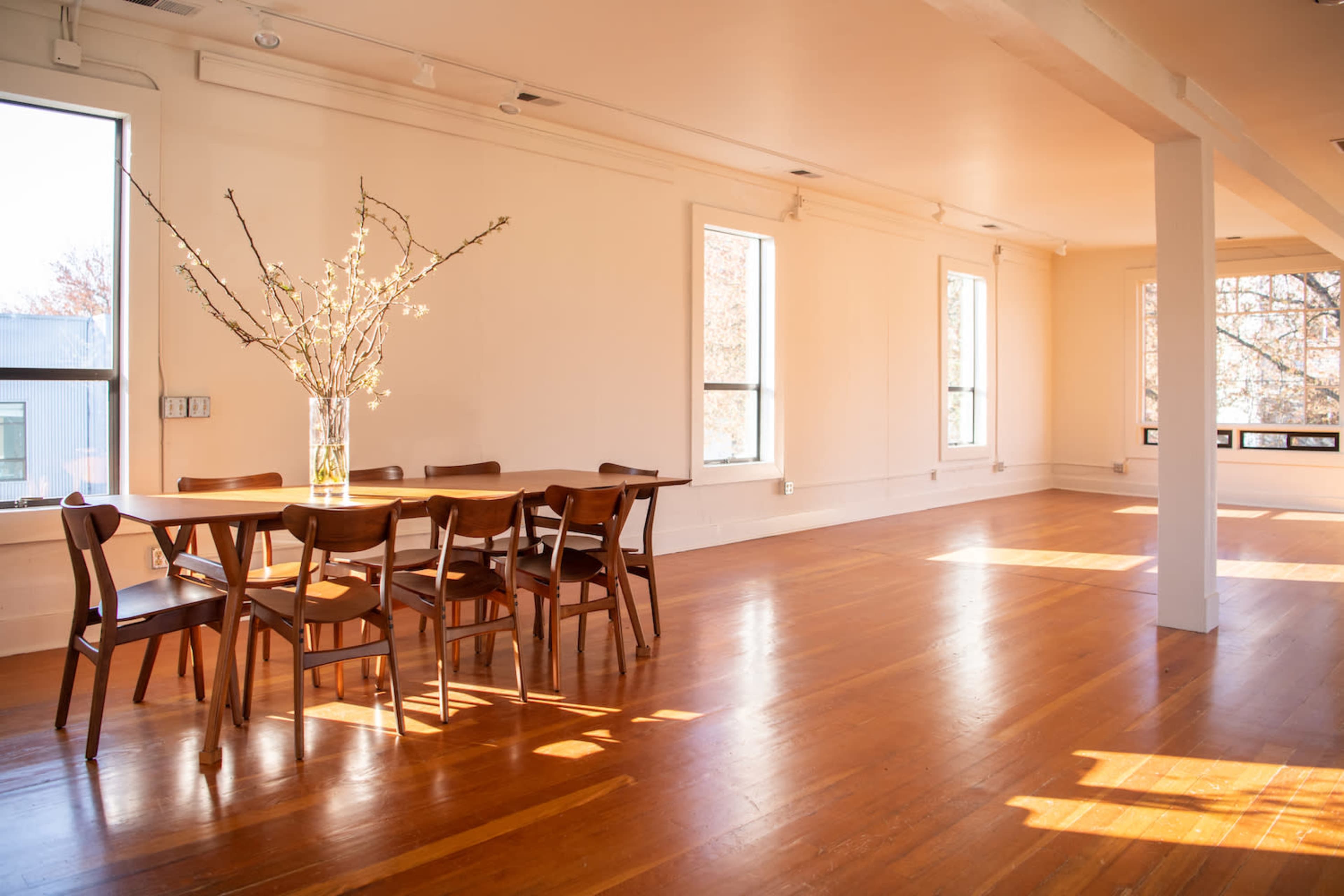 A spacious, brightly lit room features a wooden dining table surrounded by chairs, with large windows allowing sunlight to illuminate the hardwood floor.