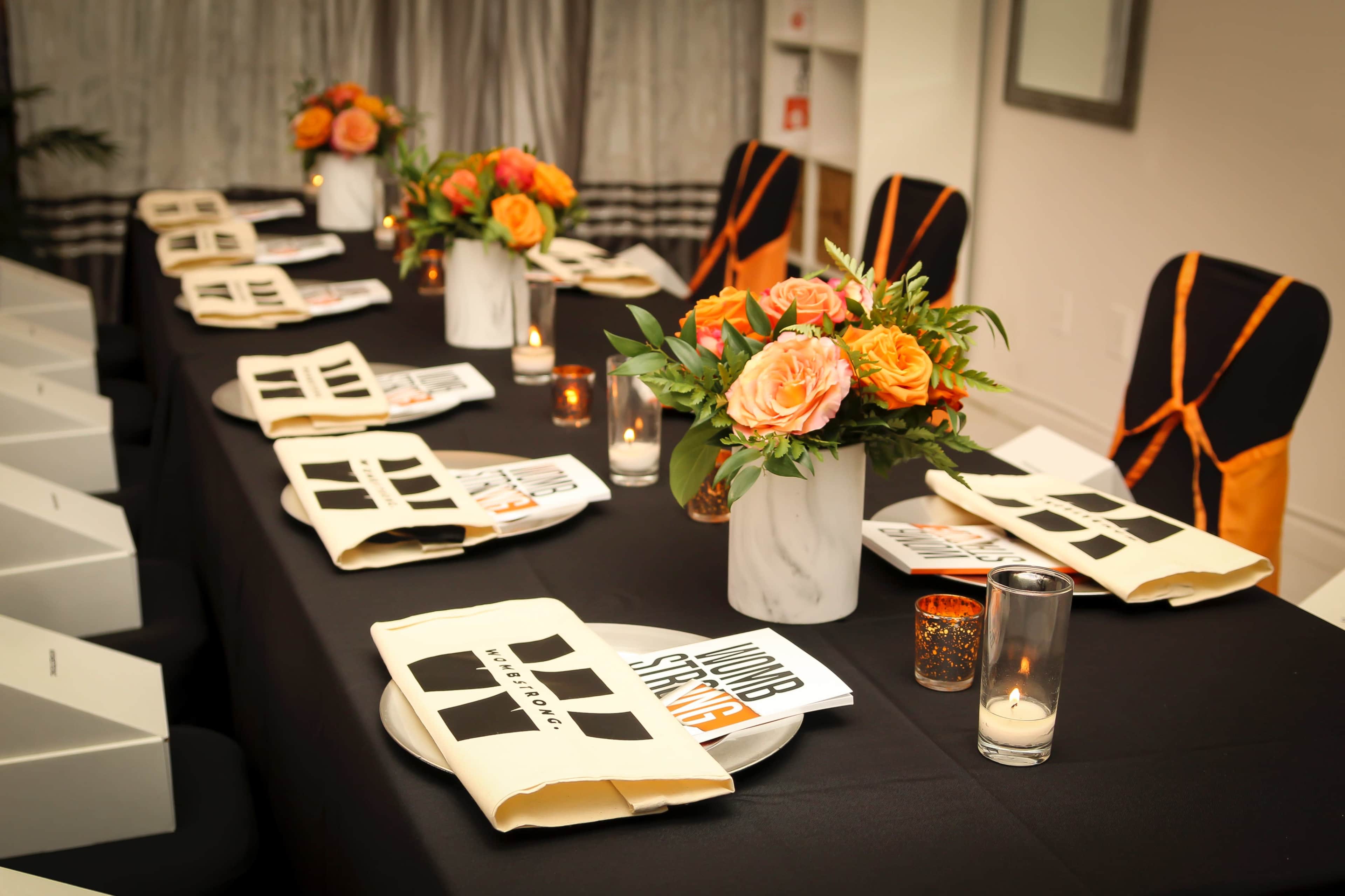 A dining table is elegantly set with black tablecloths, folded napkins, flower arrangements, and candles, ready for a formal gathering.