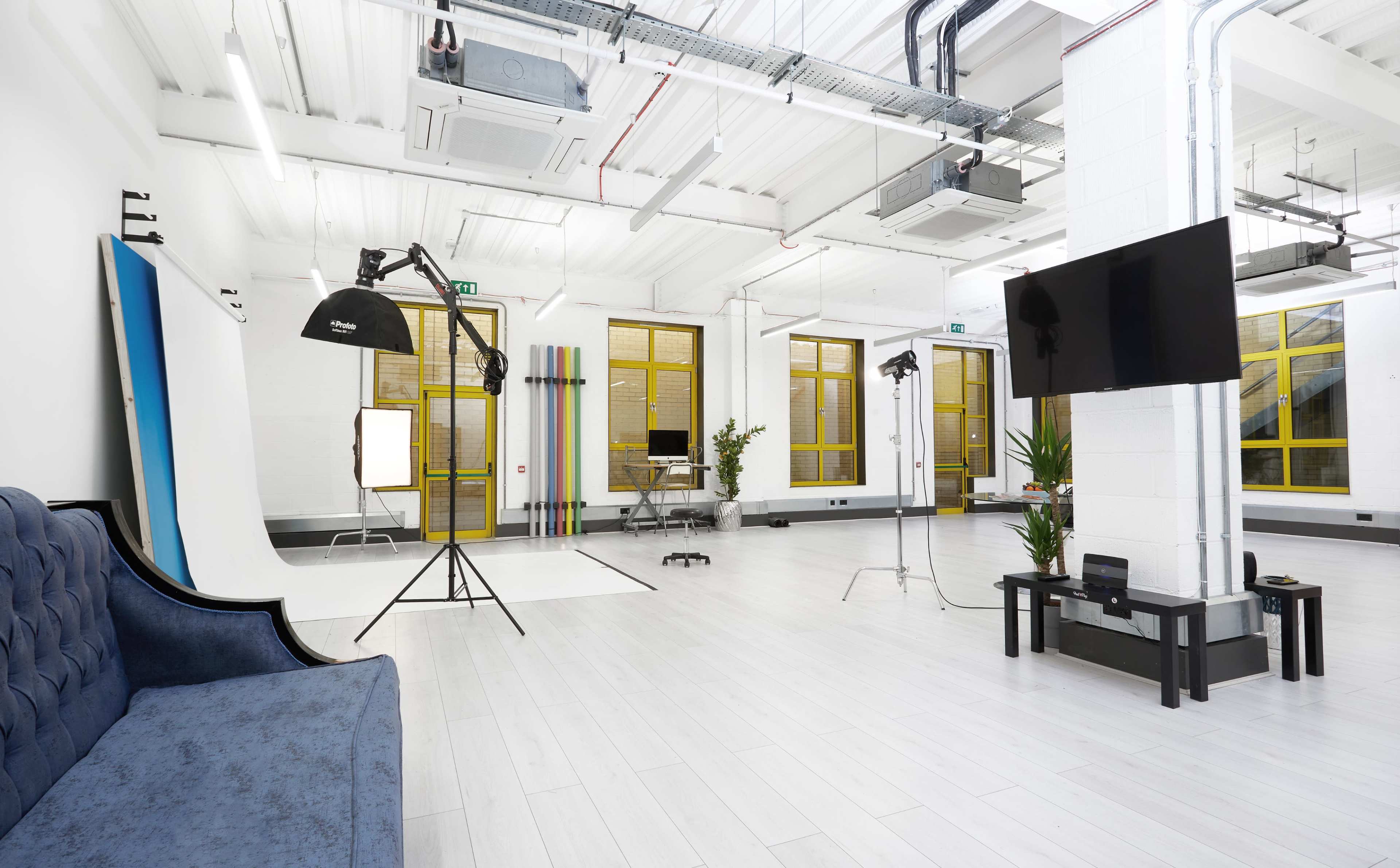 A spacious photography studio featuring a blue couch, backdrop stands, lighting equipment, and a large television mounted on the wall.