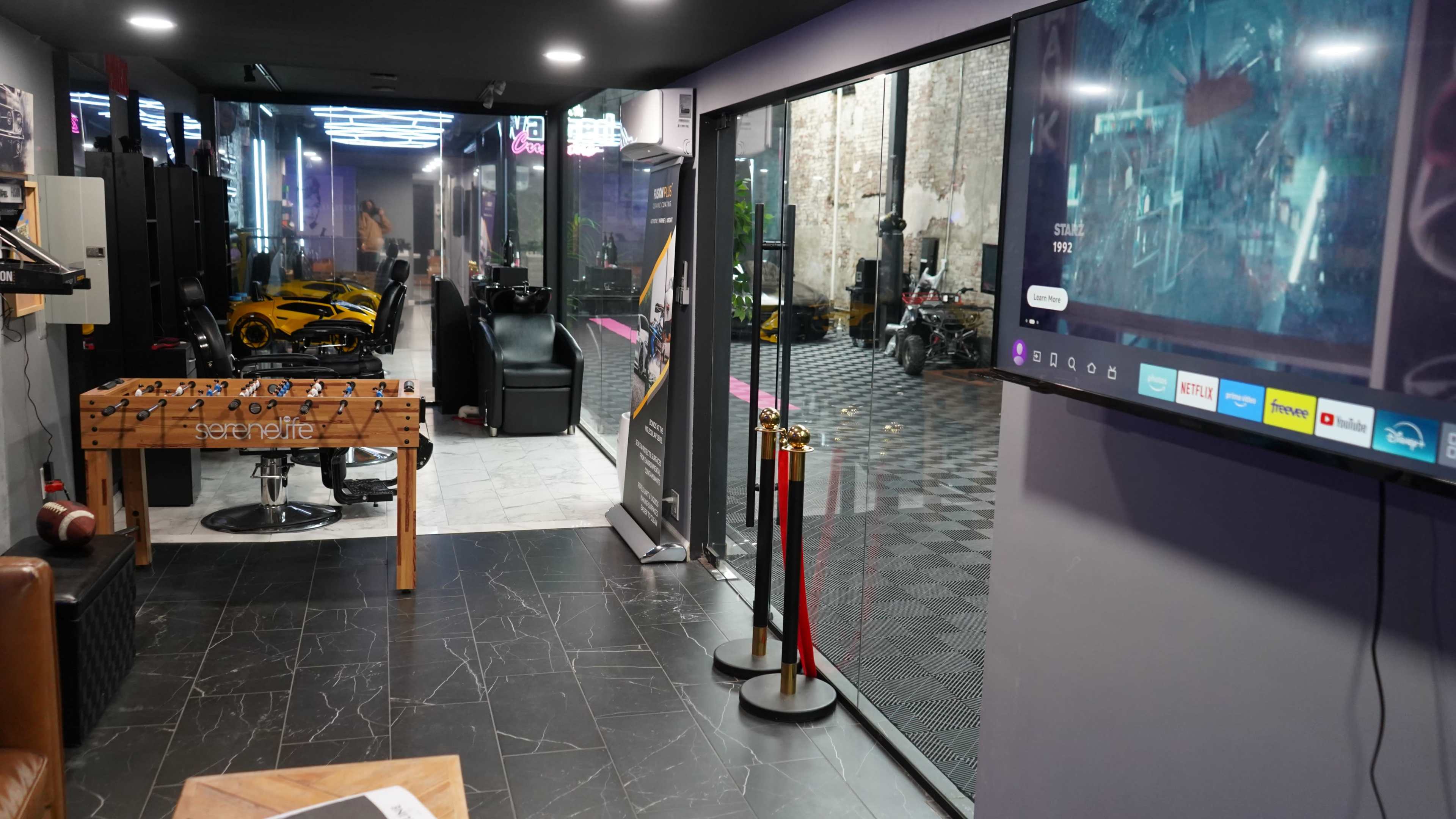 The image shows a modern indoor recreational space featuring a foosball table, a large screen displaying information, and a view of a lounge area with sleek seating and cars in the background.