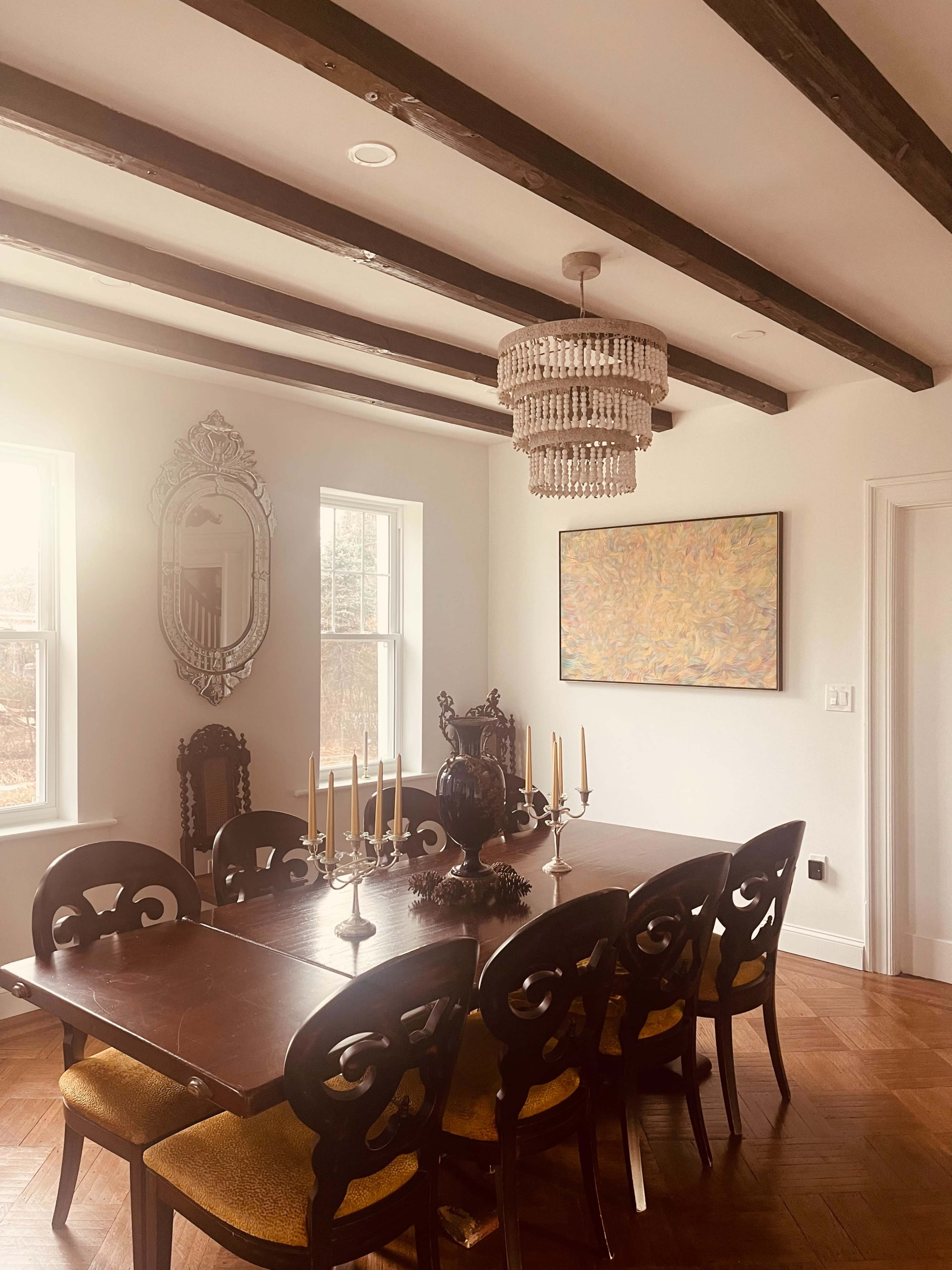 A dining room features a wooden table set for dinner, with a chandelier above and a colorful painting on the wall.