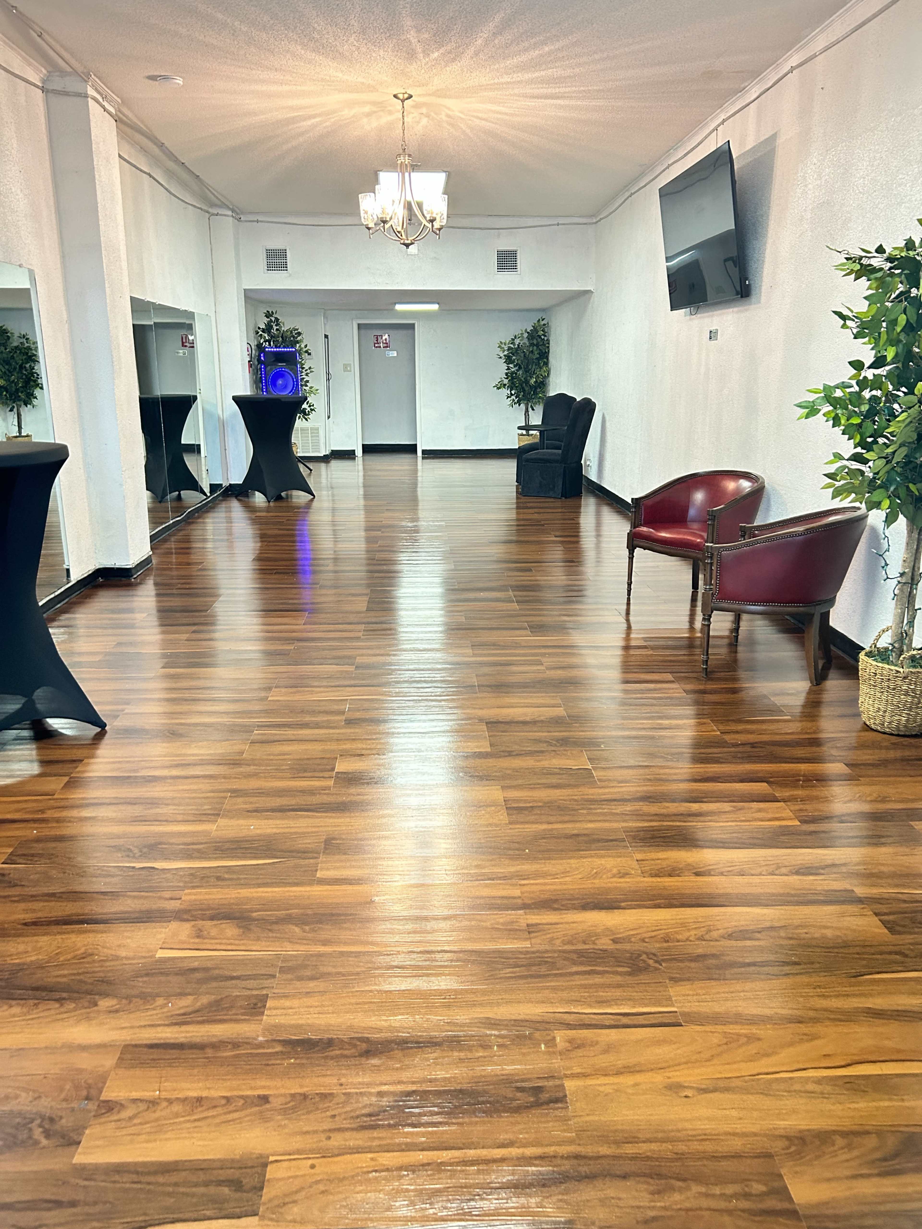 A spacious, well-lit room with polished wooden flooring, featuring a chandelier, mirrors along the walls, and a mix of seating arrangements including a chair and black cocktail tables.