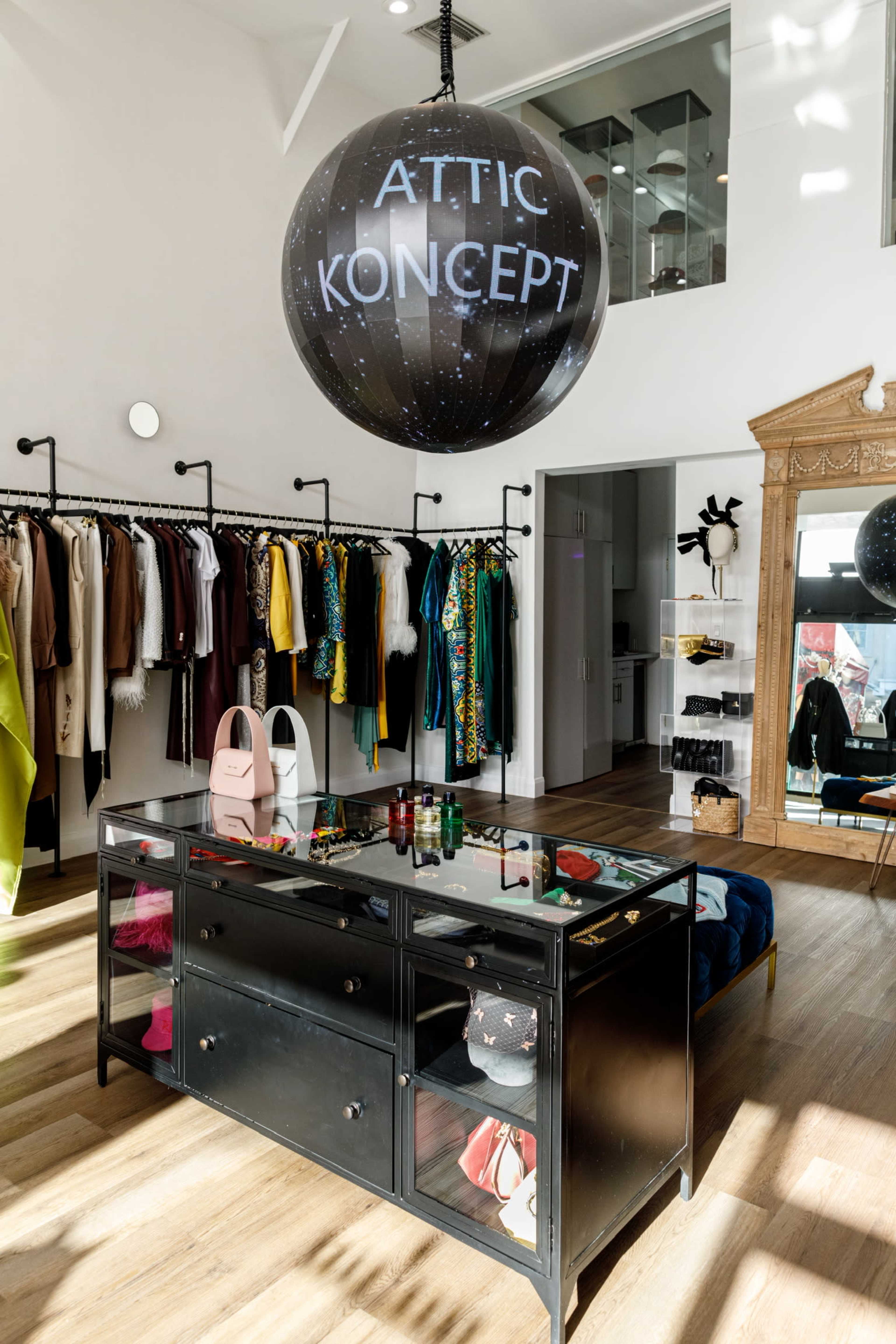 The image shows an interior of a boutique with a large black spherical sign hanging from the ceiling, displaying the text "ATTIC KONCEPT," alongside racks of colorful clothing and a mirrored display table in the center.