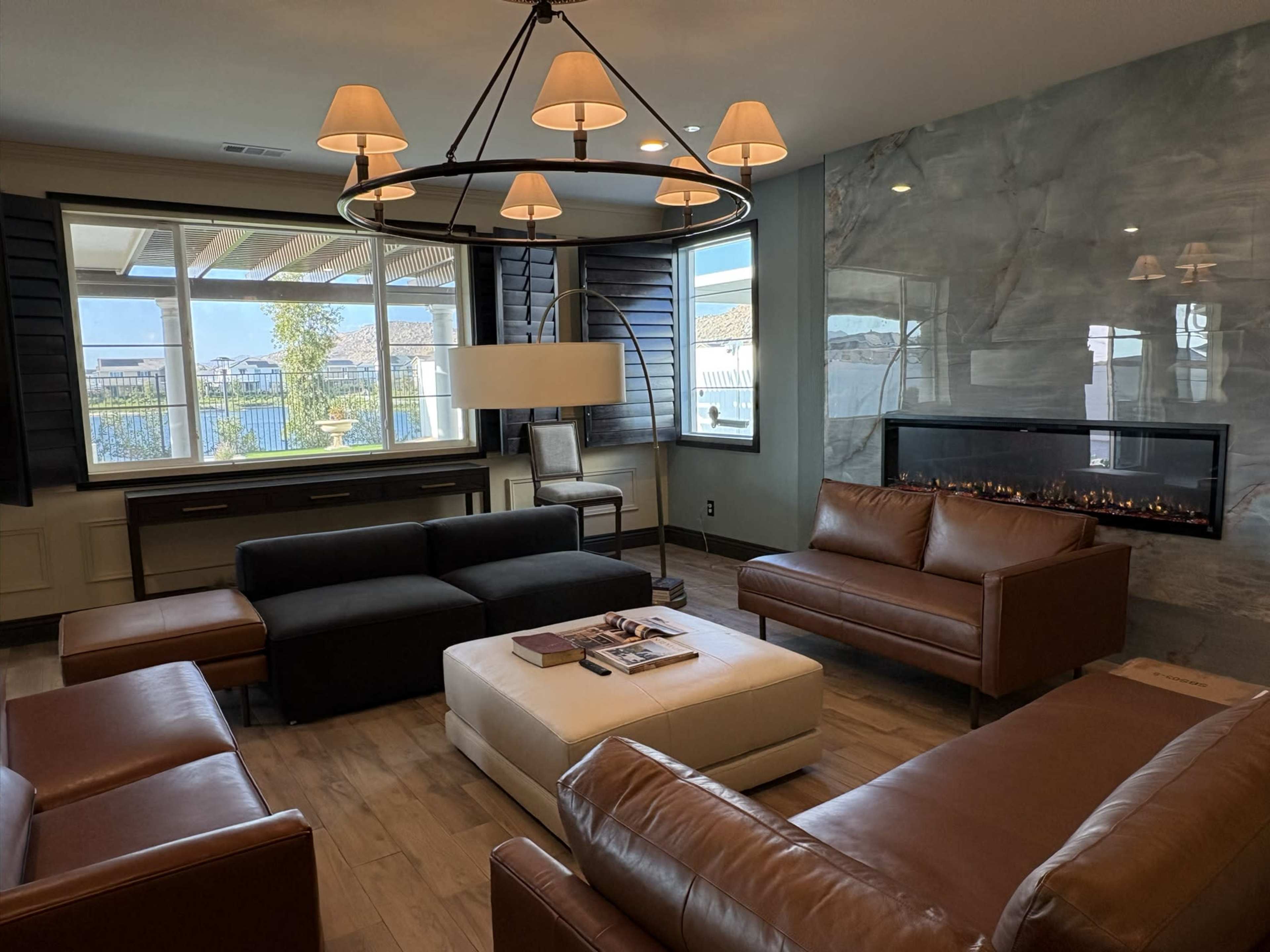 The image shows a modern living room featuring a combination of brown leather sofas, a white ottoman, a stylish coffee table, and a wall-mounted fireplace, all arranged under a decorative light fixture.