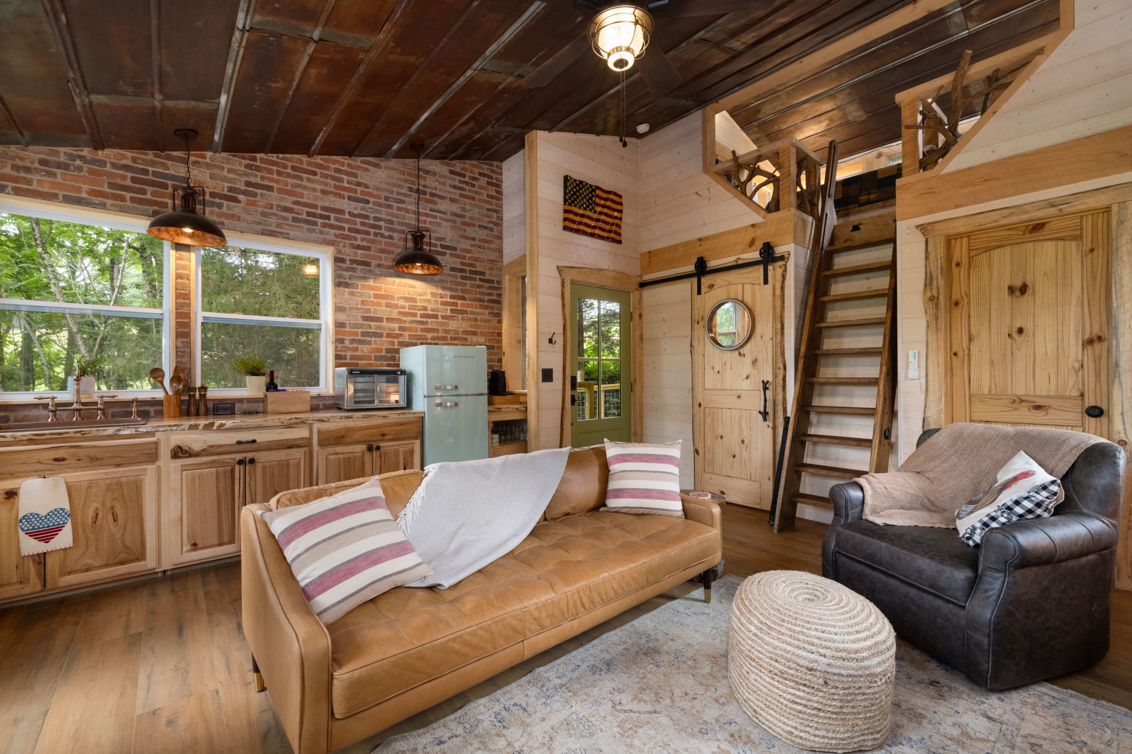 The image shows a cozy living space featuring a leather sofa, a woven pouf, and a staircase leading to an upstairs area, with exposed brick walls and wooden accents.