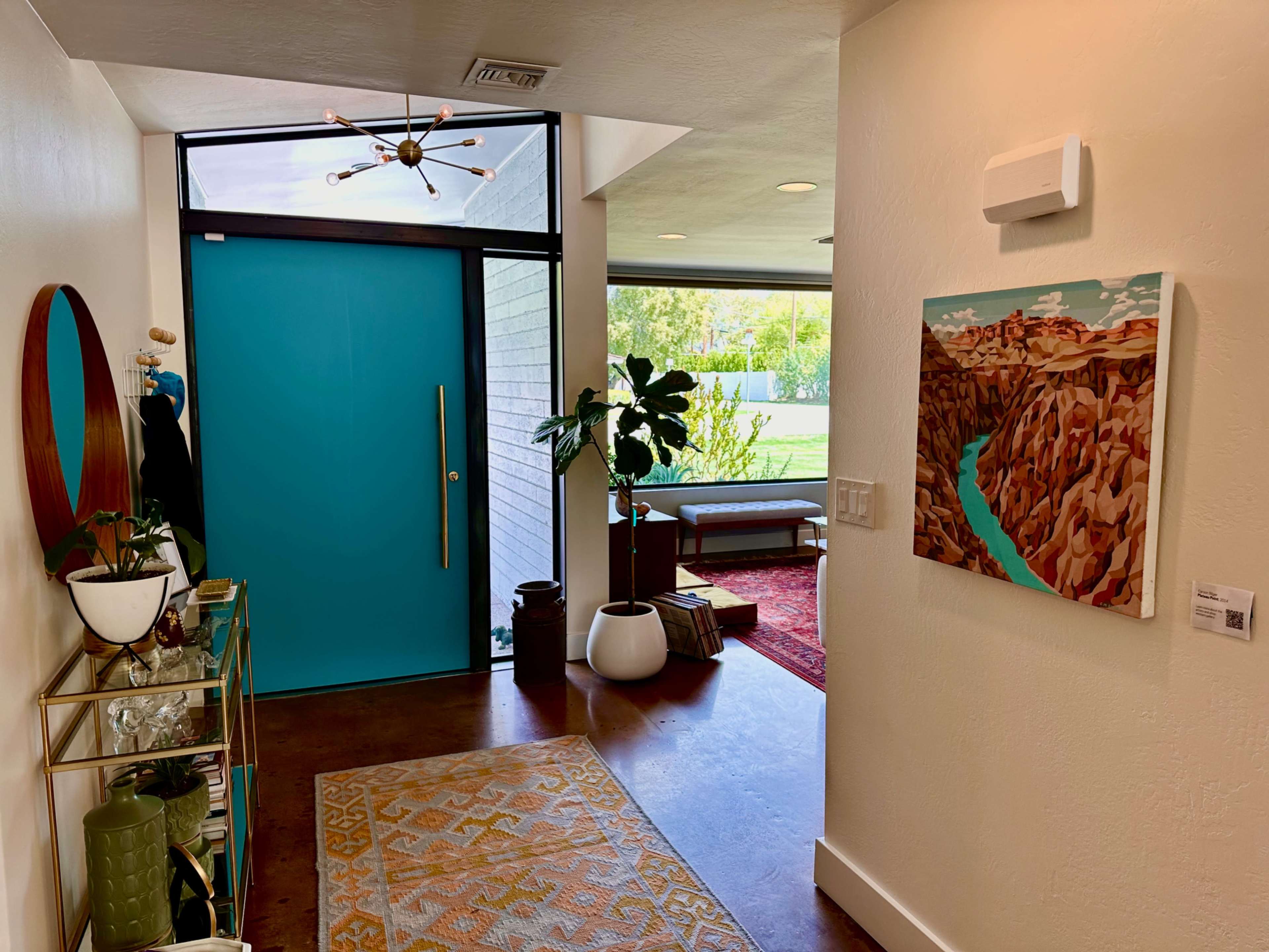 A bright entryway featuring a turquoise door, a modern console table with decorative items, and a view of a landscaped outdoor area through large windows.