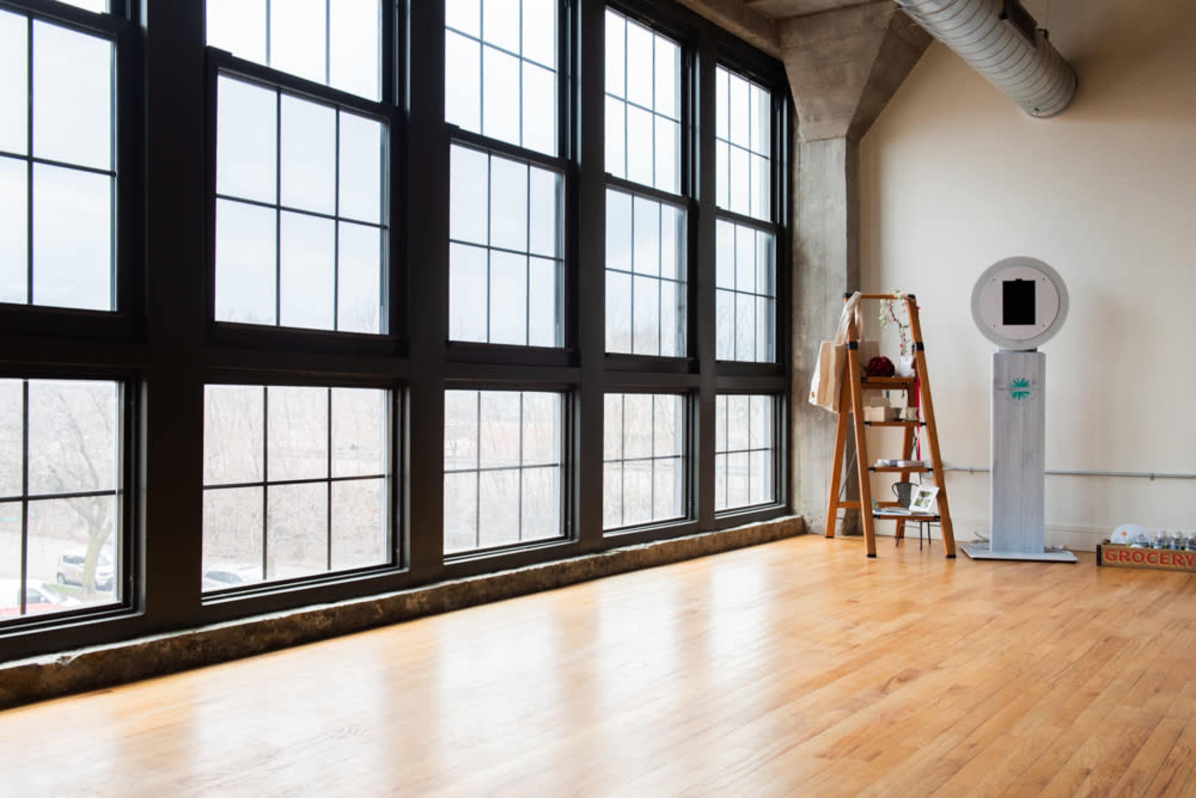 A spacious room with large windows, a wooden floor, a ladder, and a white structure resembling a kiosk.