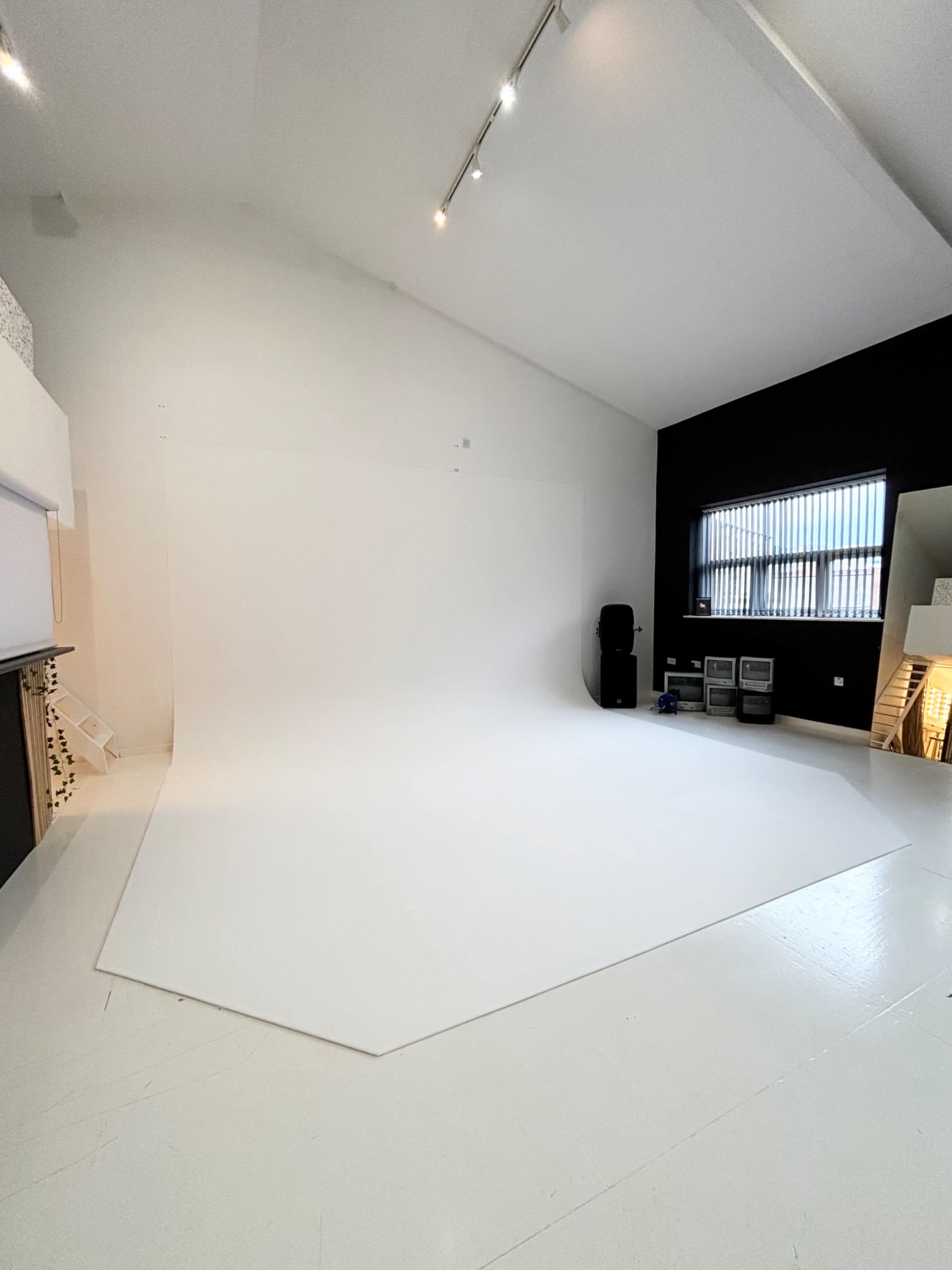 A spacious photography studio with a large white backdrop and a black wall, equipped with various studio equipment in the corner.