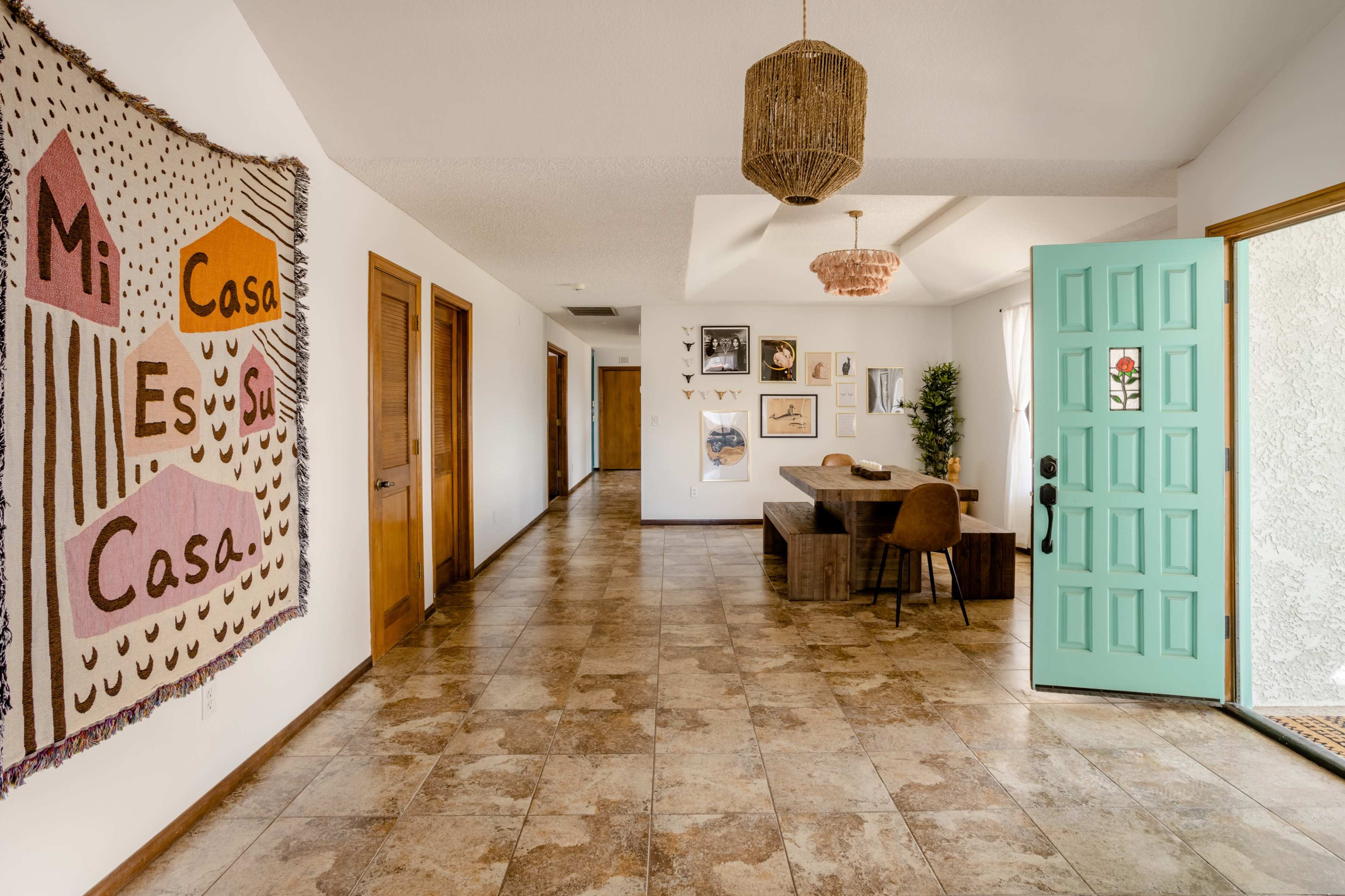 A brightly lit entryway features a woven wall hanging, a turquoise door, and a wooden dining table beside a seating area.