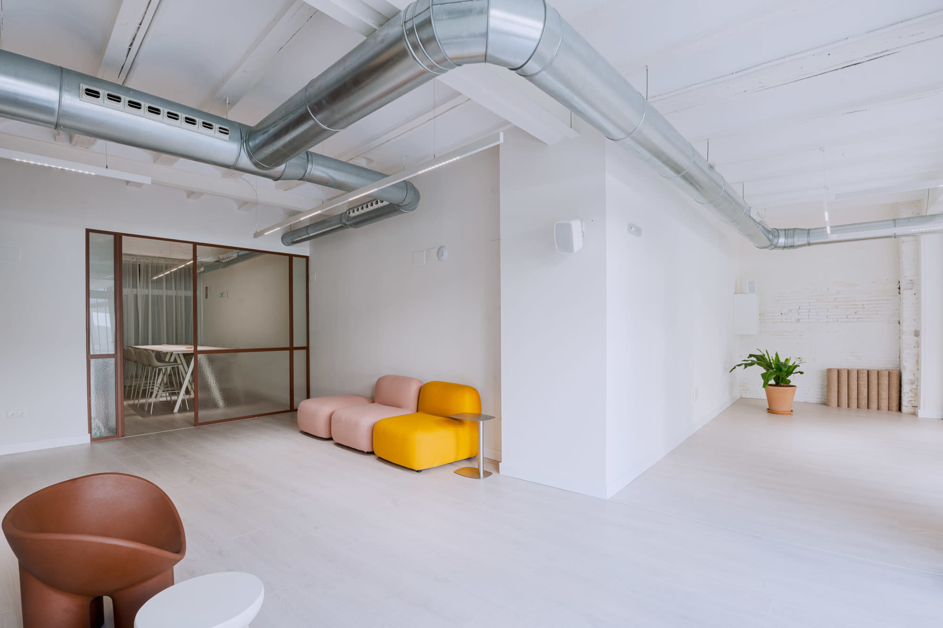 The image shows a modern, open office space featuring two seating areas, large windows, and exposed ductwork on the ceiling.