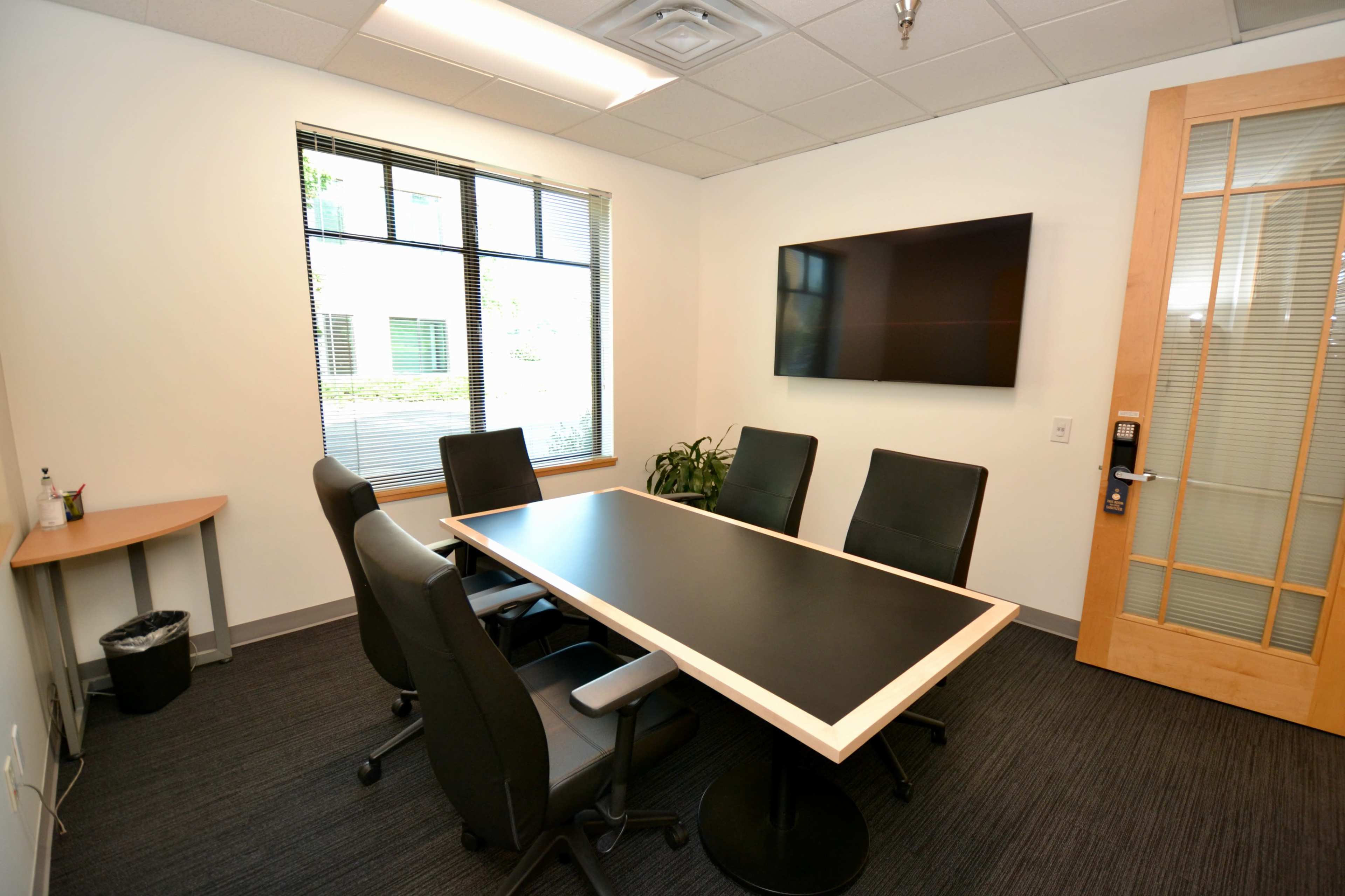A small conference room with a black table, five chairs, a television on the wall, and a window with blinds.