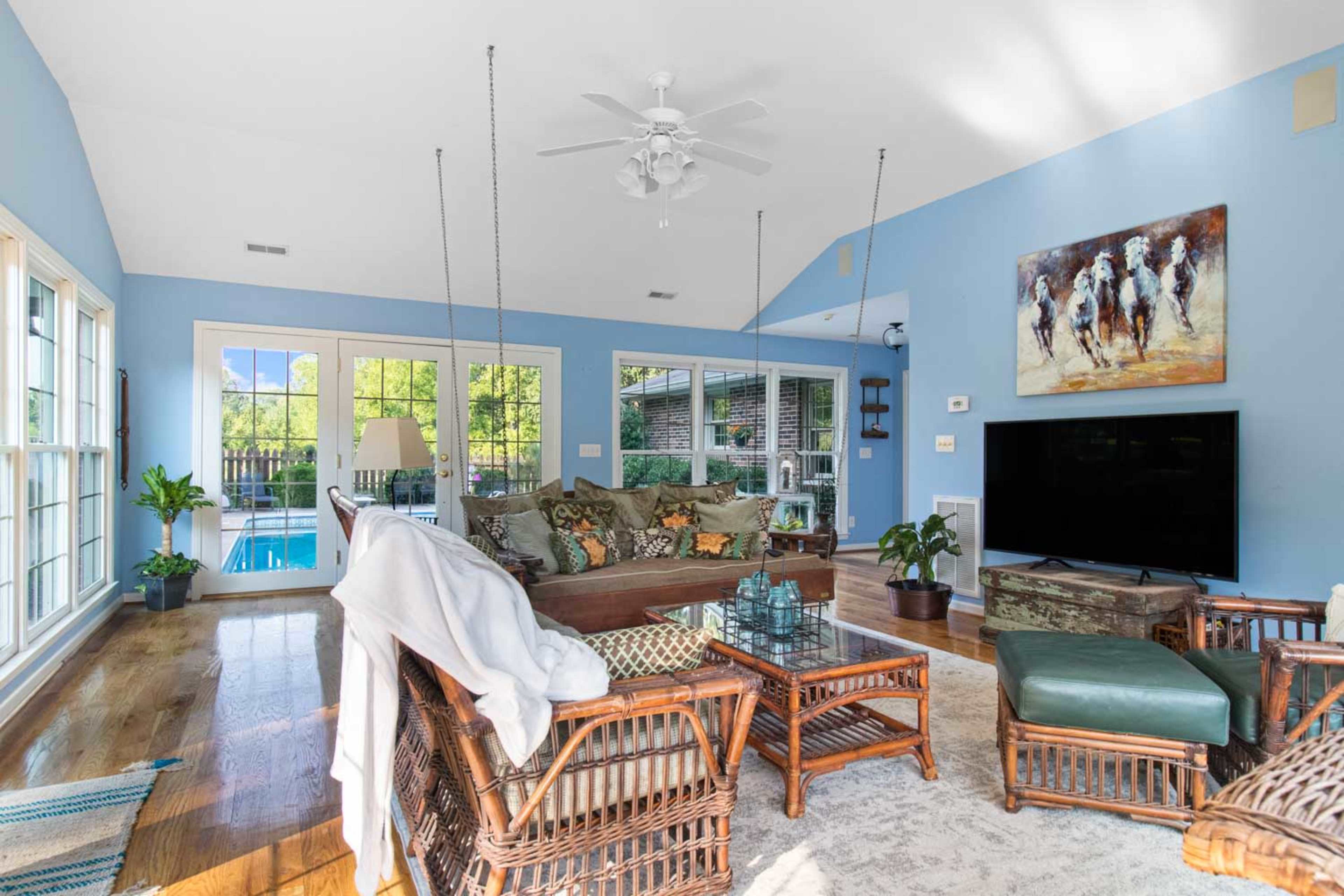 A sunlit living room with blue walls, a large sofa, a coffee table, and a television, with a view of a pool through the windows.