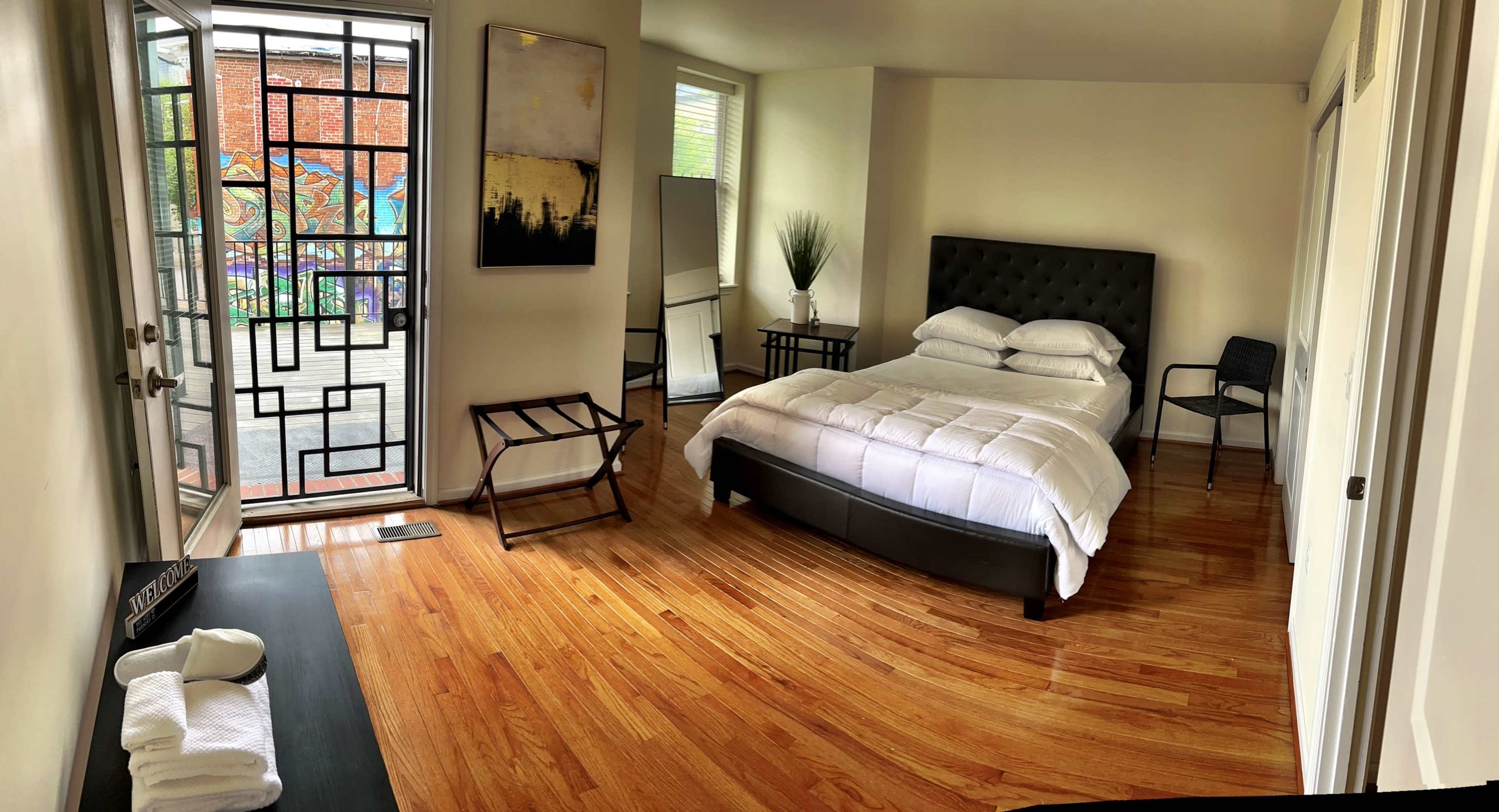 A neatly arranged bedroom features a bed with white linens, wooden floors, and a door leading to a patio area with artwork visible outside.