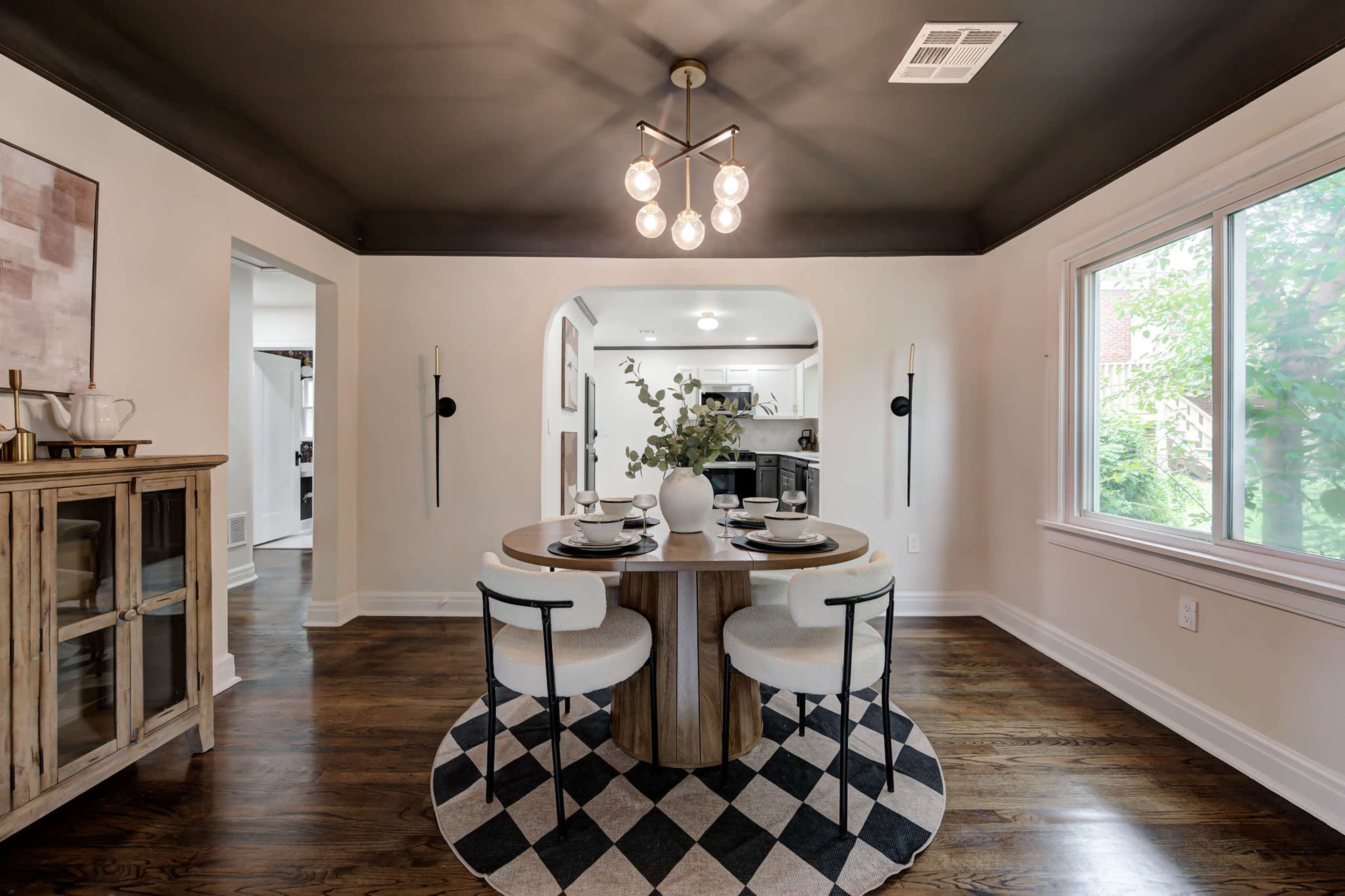A modern dining area features a round table with white chairs, surrounded by a black and white geometric rug, and a large window allowing natural light to enter.