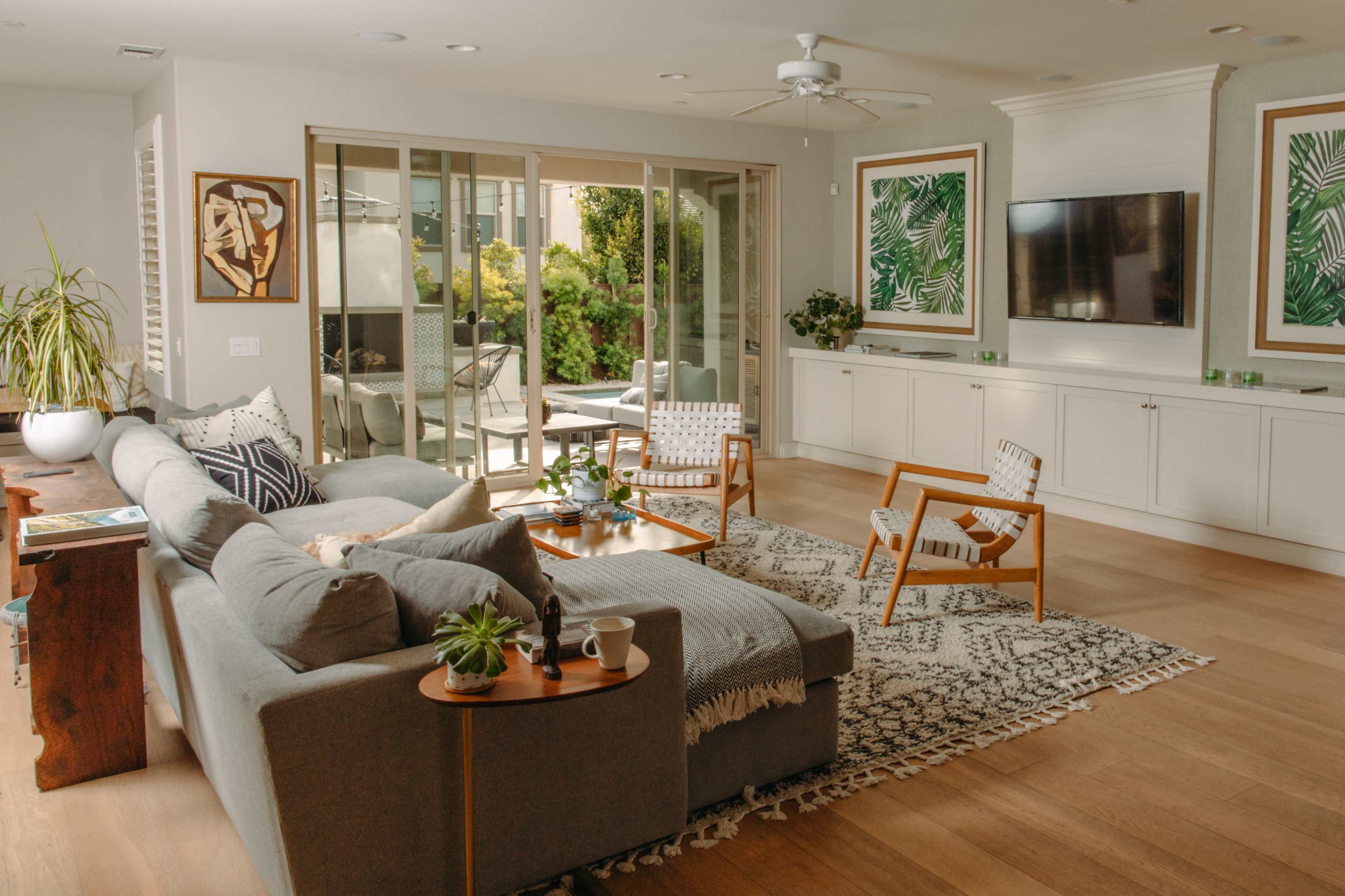 A modern living room features a gray sectional sofa, a patterned area rug, wooden chairs, and large windows opening to a patio with greenery.