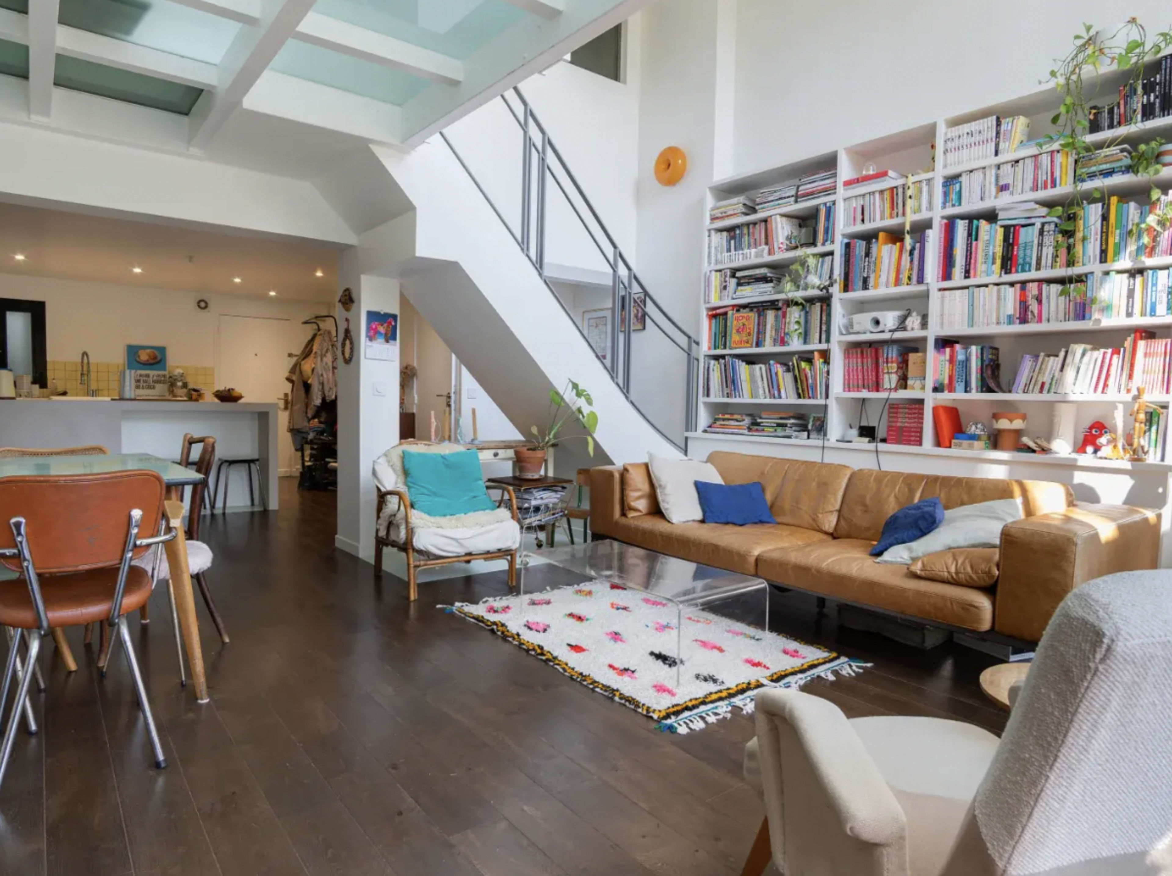 The image shows a modern, open living space featuring a brown leather sofa, a colorful rug, a large bookshelf filled with books, and a dining area with wooden chairs.