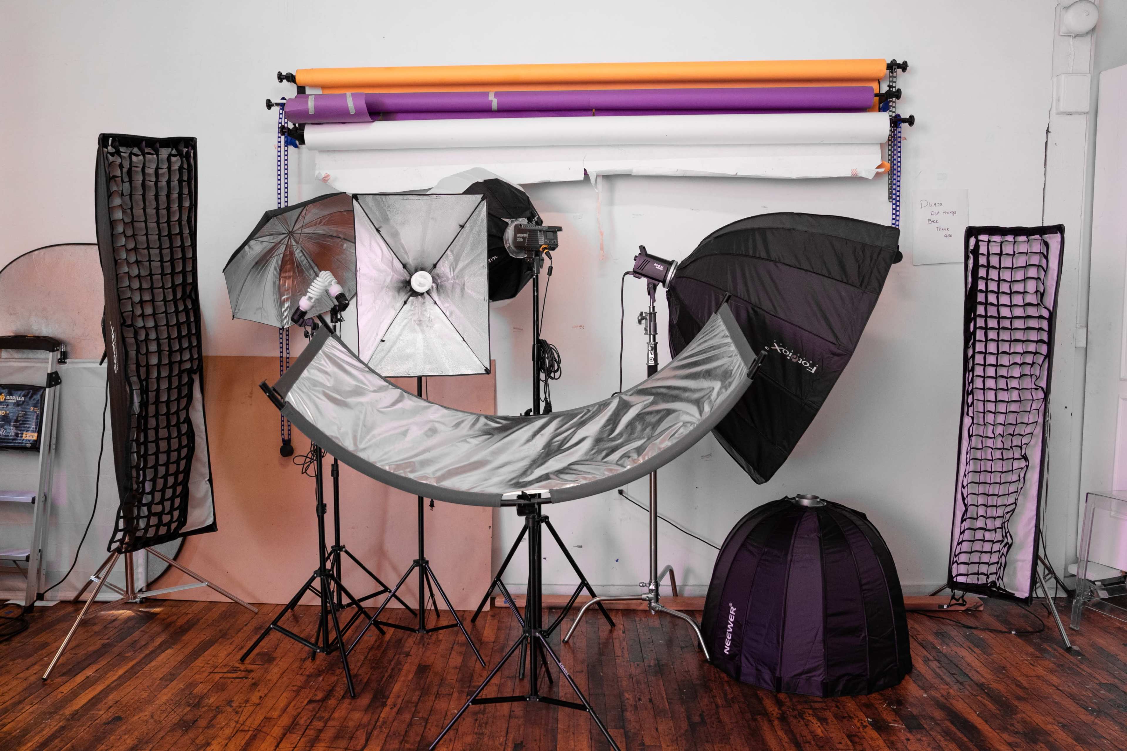 The image shows a photography studio setup with various lighting equipment, reflectors, and backdrops arranged against a wall.