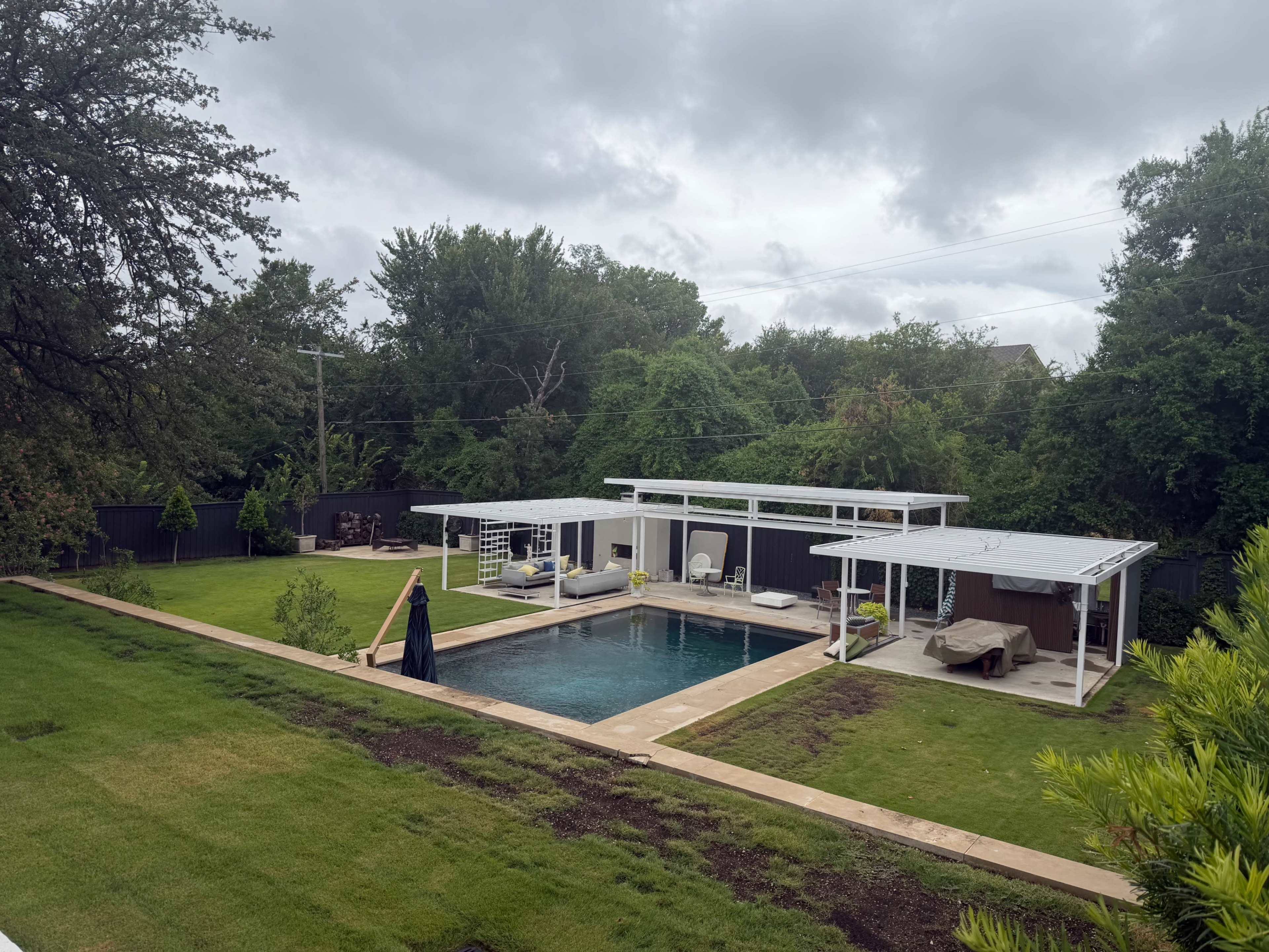 The image shows a modern outdoor pool area with two covered lounging spaces, surrounded by greenery and a neatly trimmed lawn.