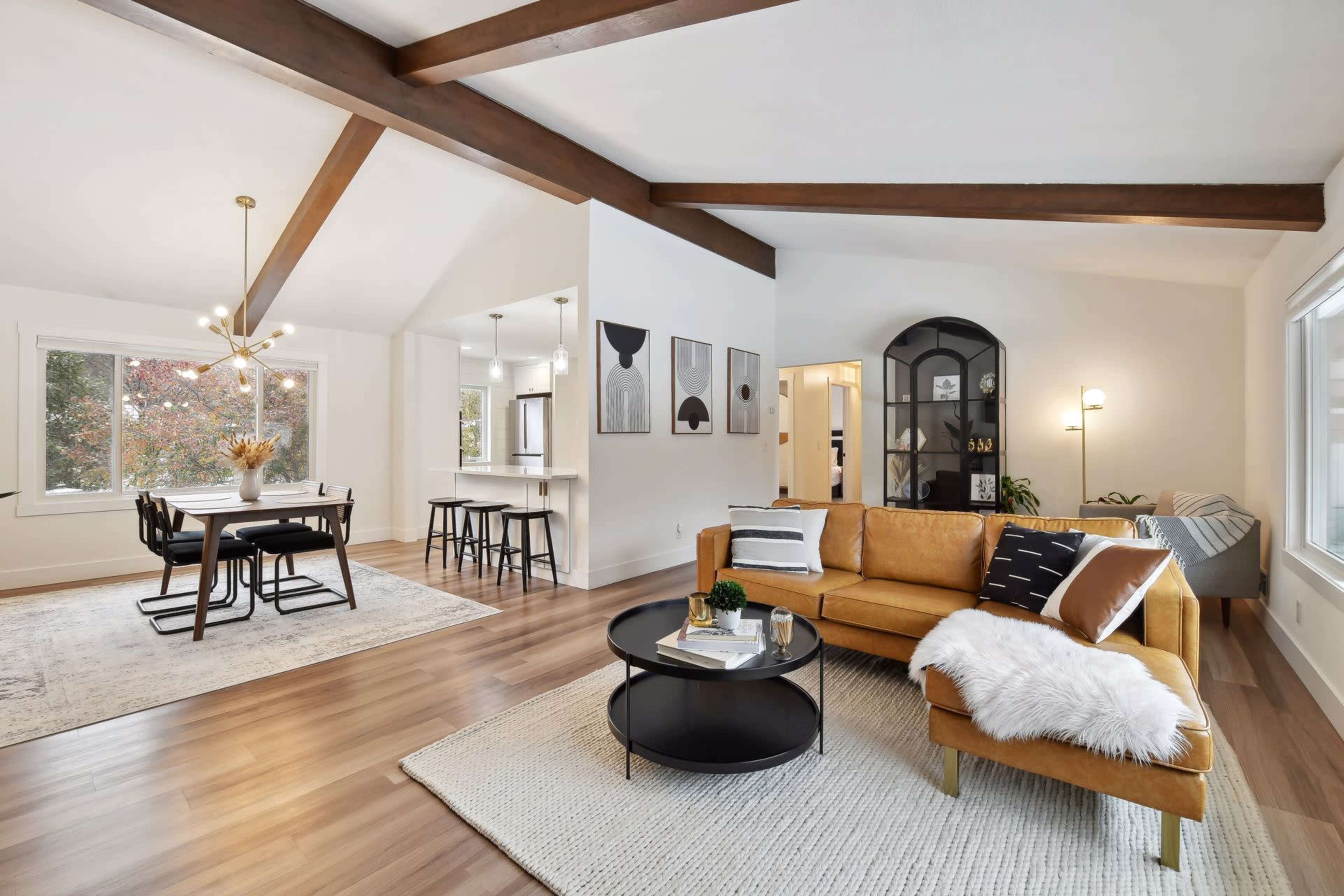 A bright, modern living room with a tan sofa, round coffee table, and wooden beams, connected to a dining area with a black table and chairs.