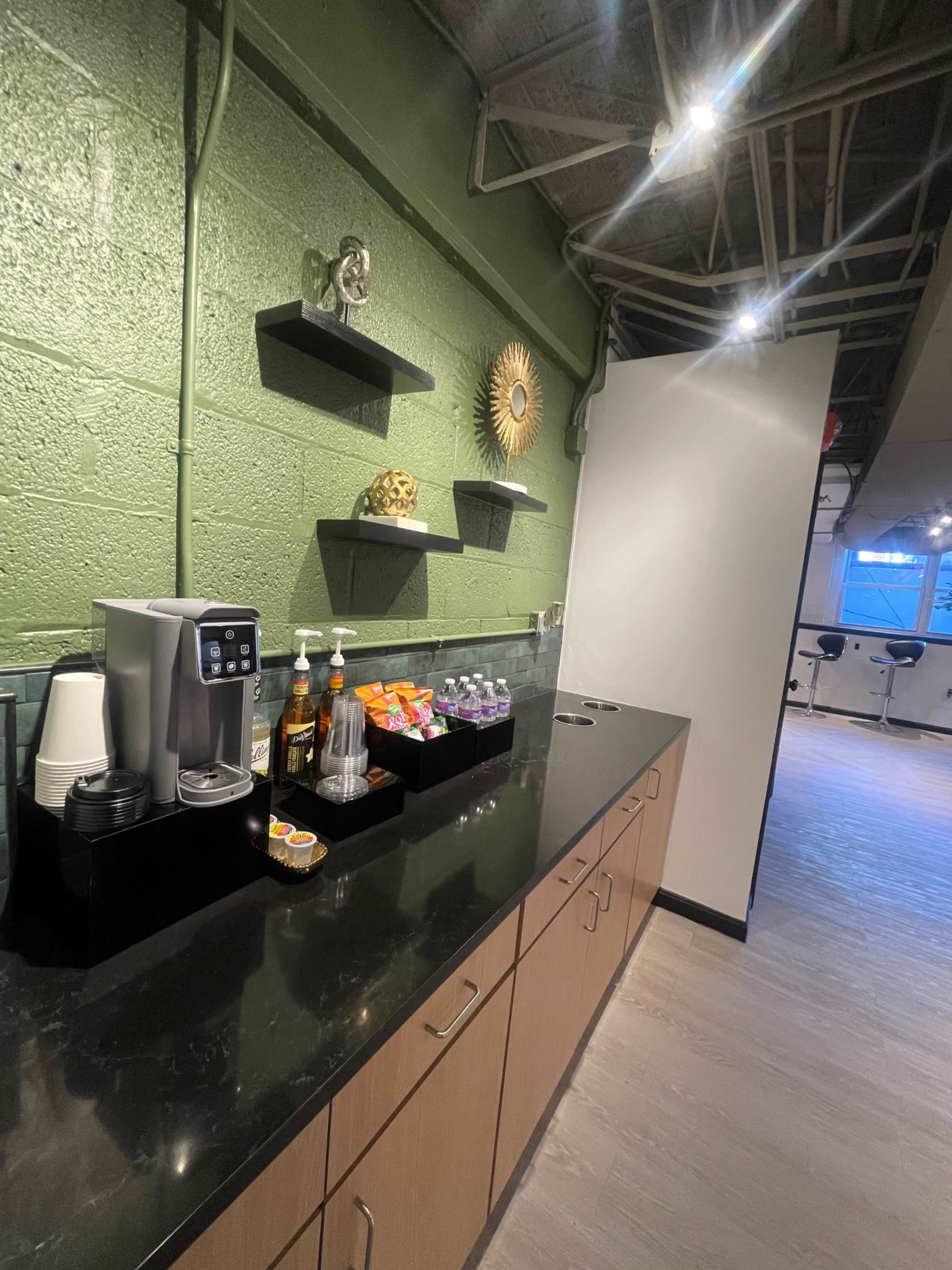 The image shows a counter with a drink station, featuring an electric beverage dispenser, various bottled drinks, and condiments, against a green wall.