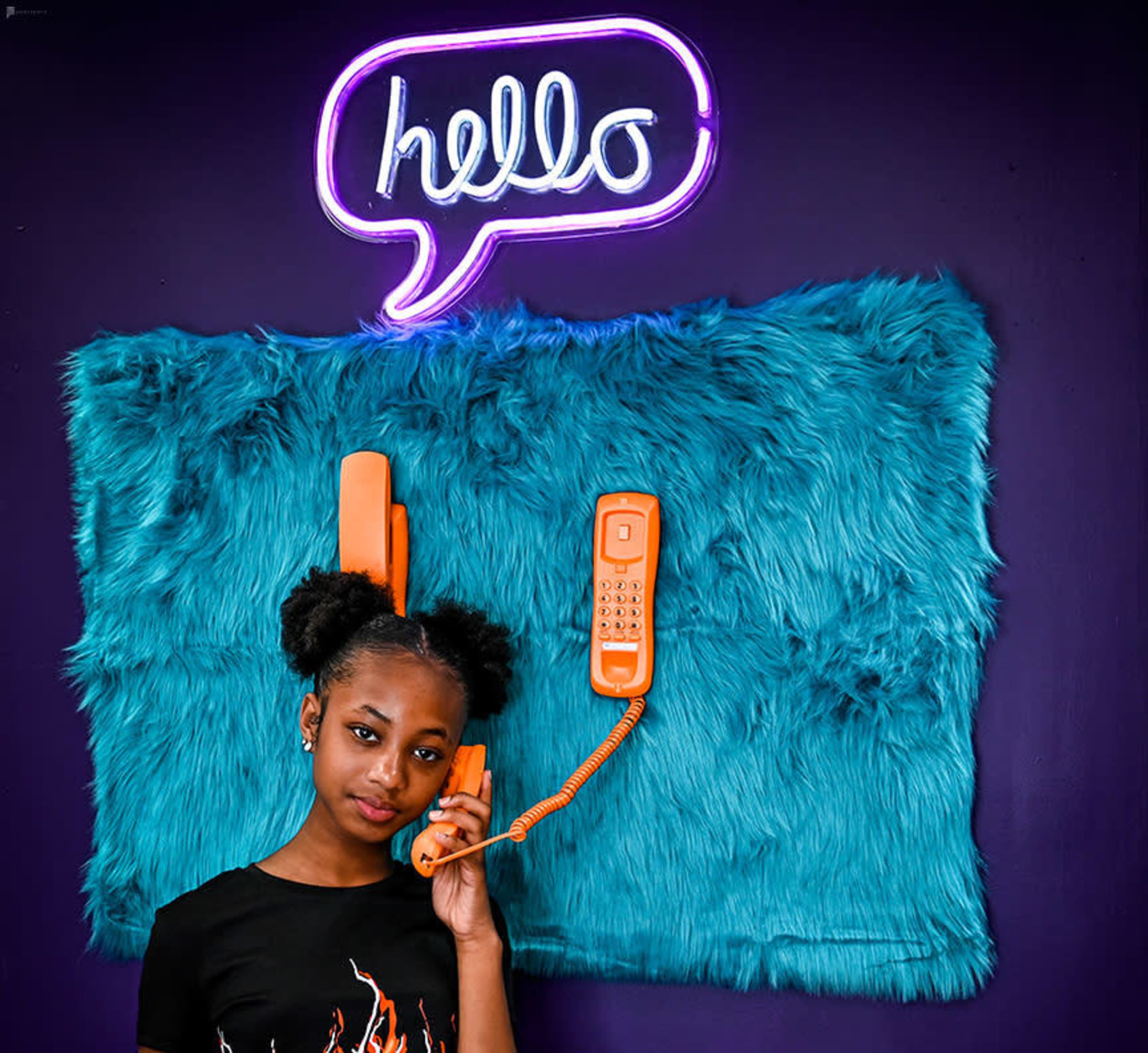 A girl with puffy hair holds an orange telephone in front of a furry blue backdrop and a neon sign that says "hello."