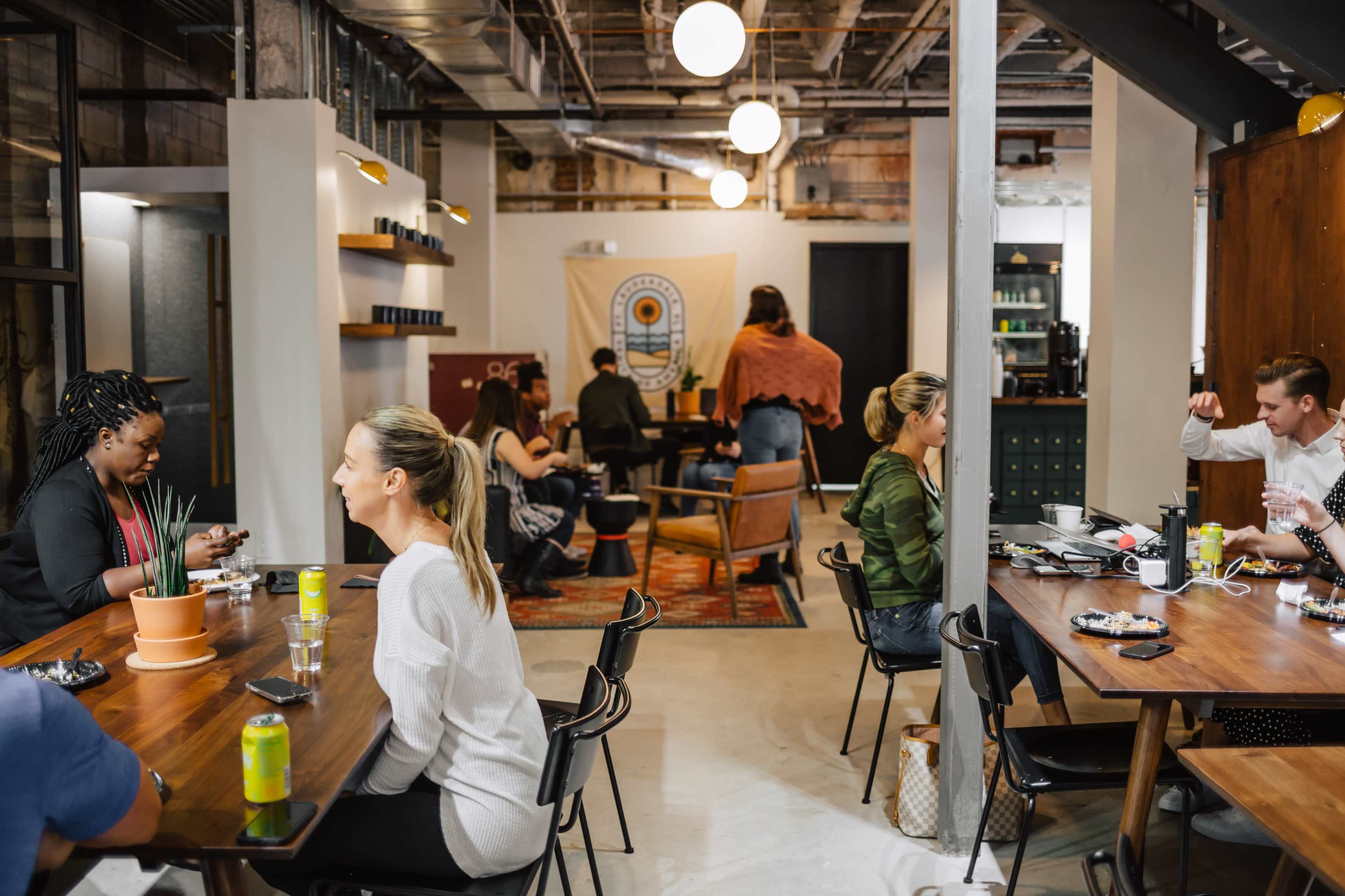 A lively cafe interior with people sitting at wooden tables, engaging in conversation and enjoying drinks, while a large decorative banner hangs in the background.