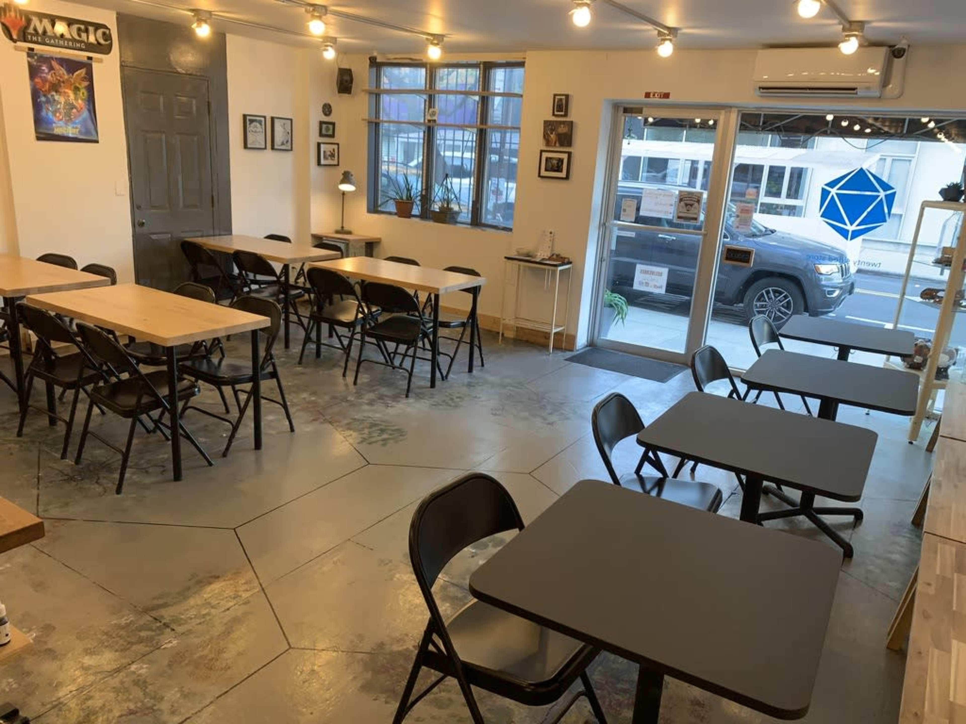 A café interior with several tables and chairs arranged neatly, large windows letting in natural light, and an entrance door leading to the street.