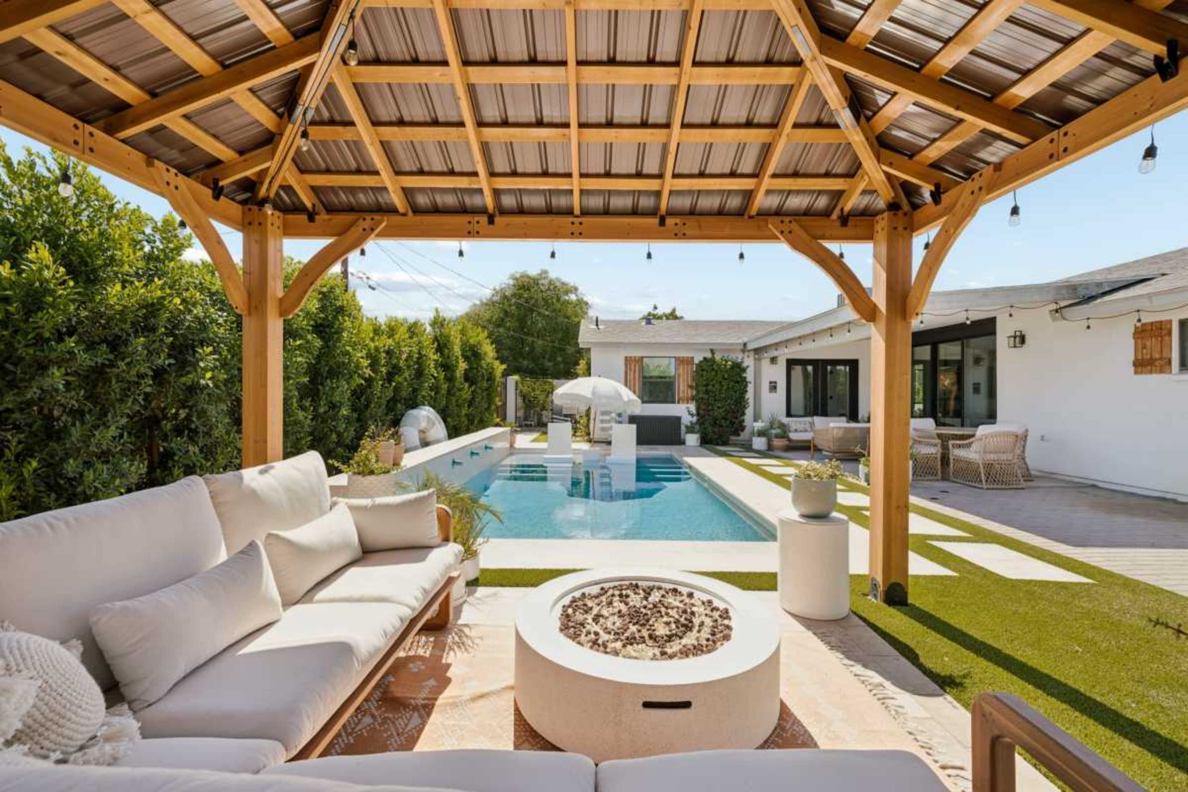 A wooden pergola shades a seating area with a fire pit, overlooking a modern pool and landscaped yard.