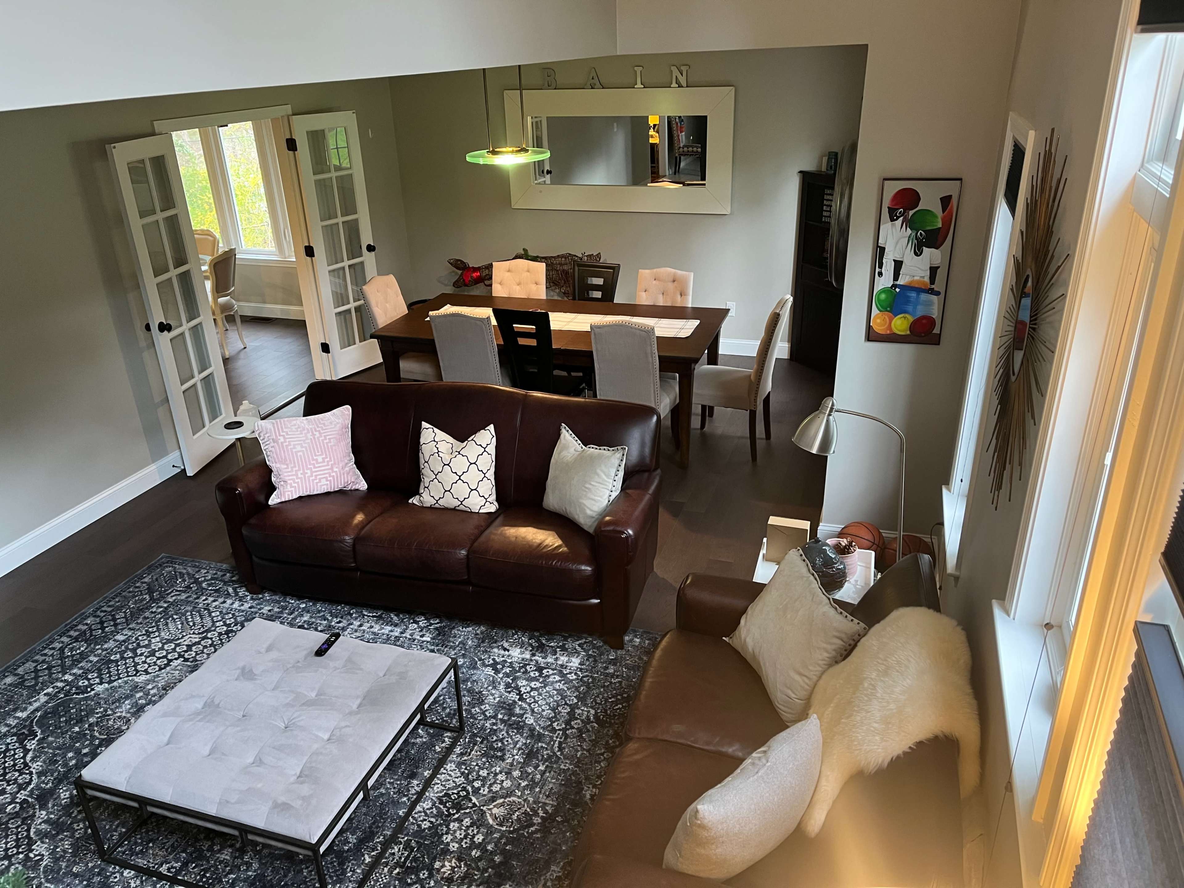 A spacious living room features a brown leather sofa, a light-colored rug, and a dining area with a wooden table set for six.