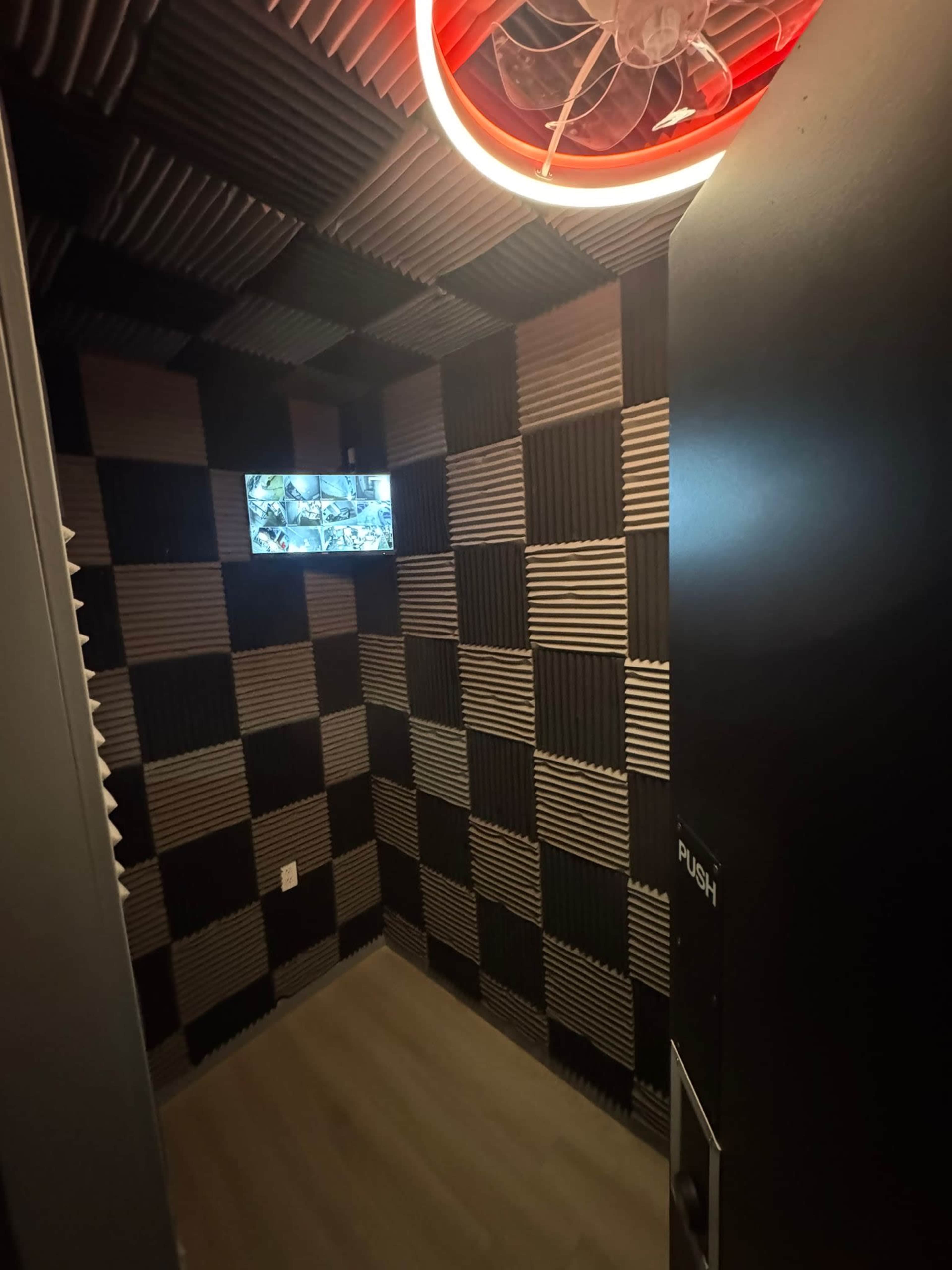 The image shows a small room with black and gray acoustical paneling on the walls, featuring a monitor on one wall and a circular light fixture on the ceiling.