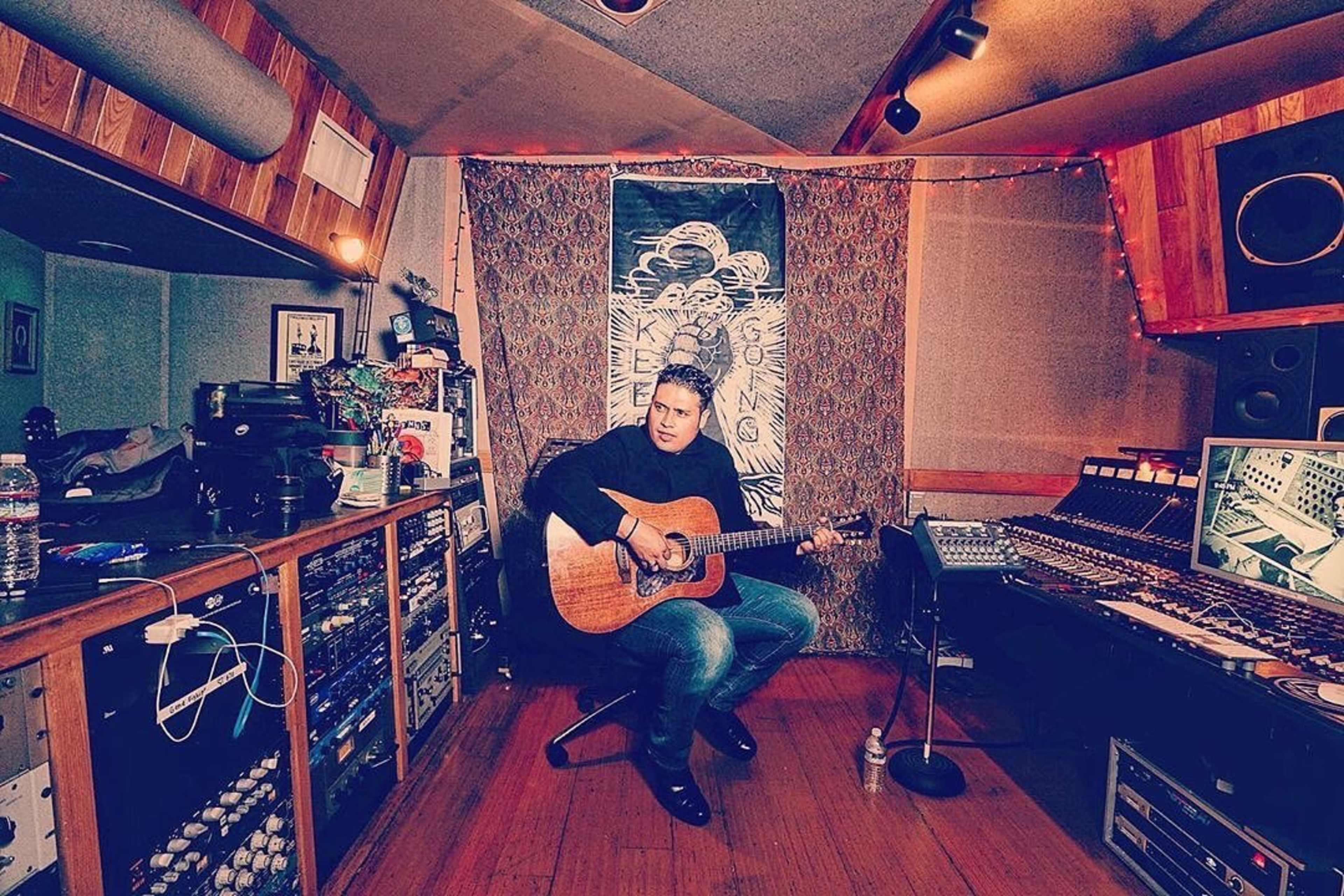 A musician sits on the floor of a recording studio, playing an acoustic guitar surrounded by audio equipment and a colorful backdrop.