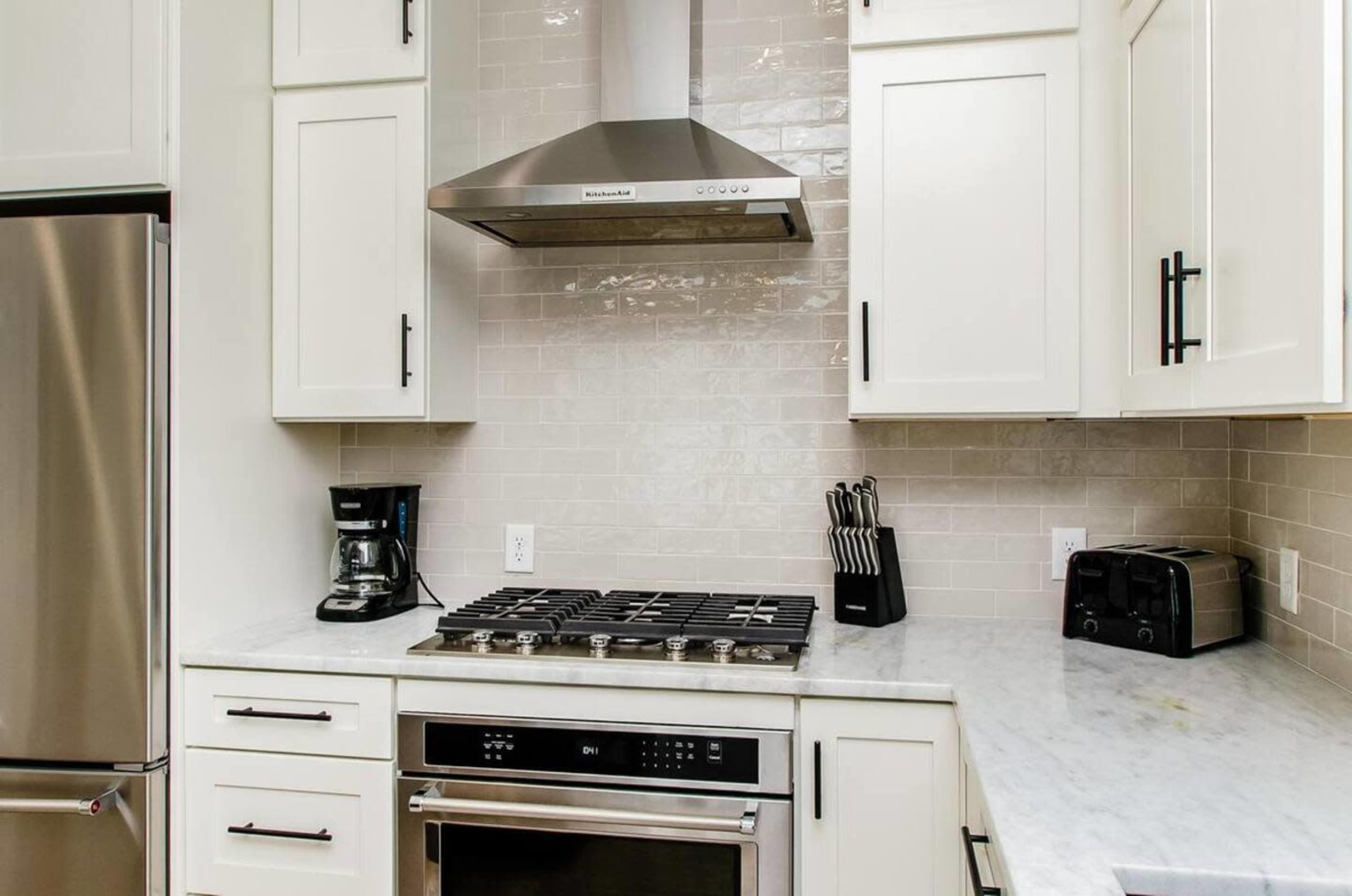 The image shows a modern kitchen with white cabinetry, a stainless steel refrigerator, a coffee maker, and a gas stove with a hood.