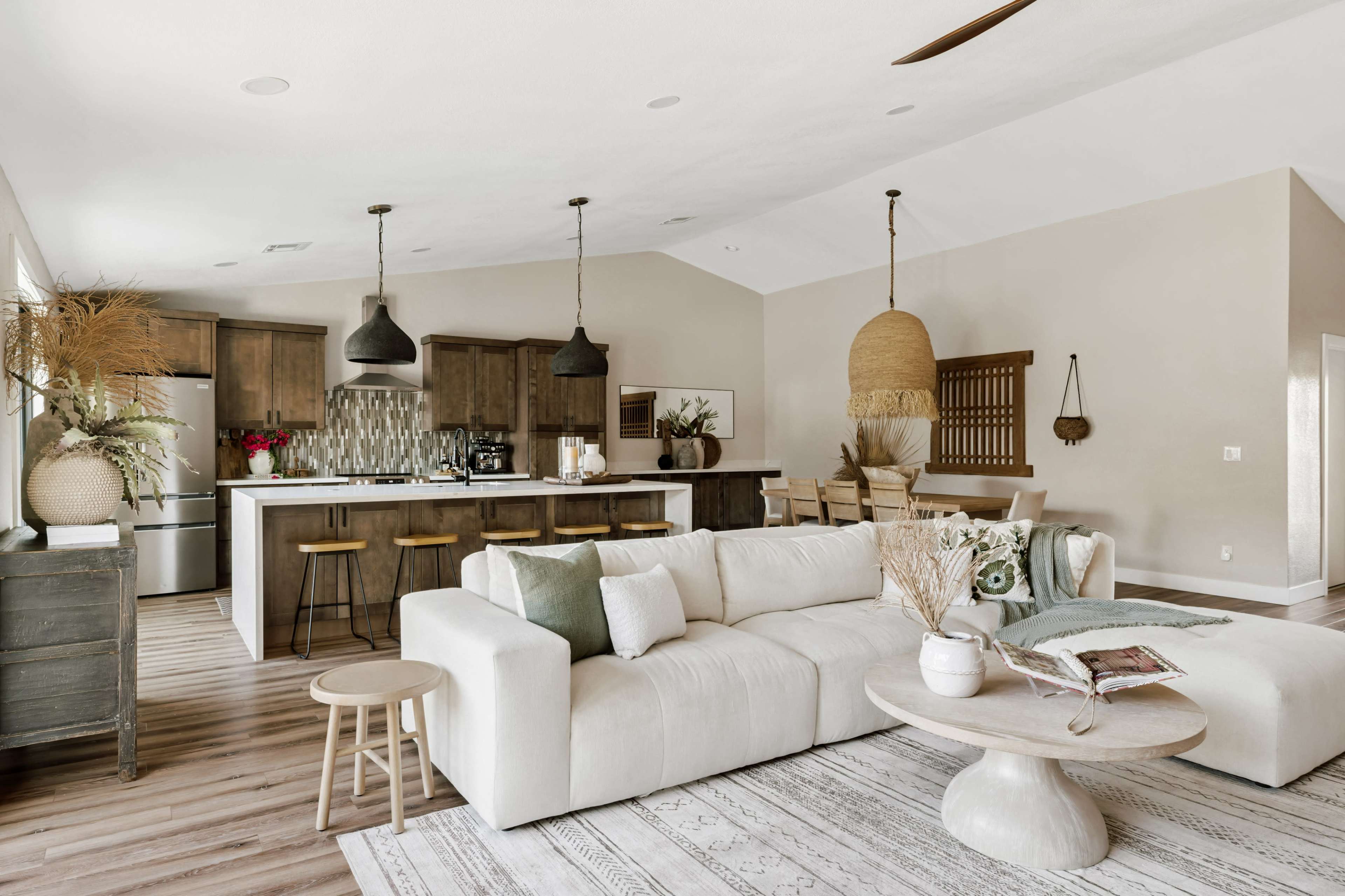 The image shows a modern living room with a sectional sofa, a round coffee table, and a kitchen area in the background featuring dark wooden cabinetry and pendant lighting.