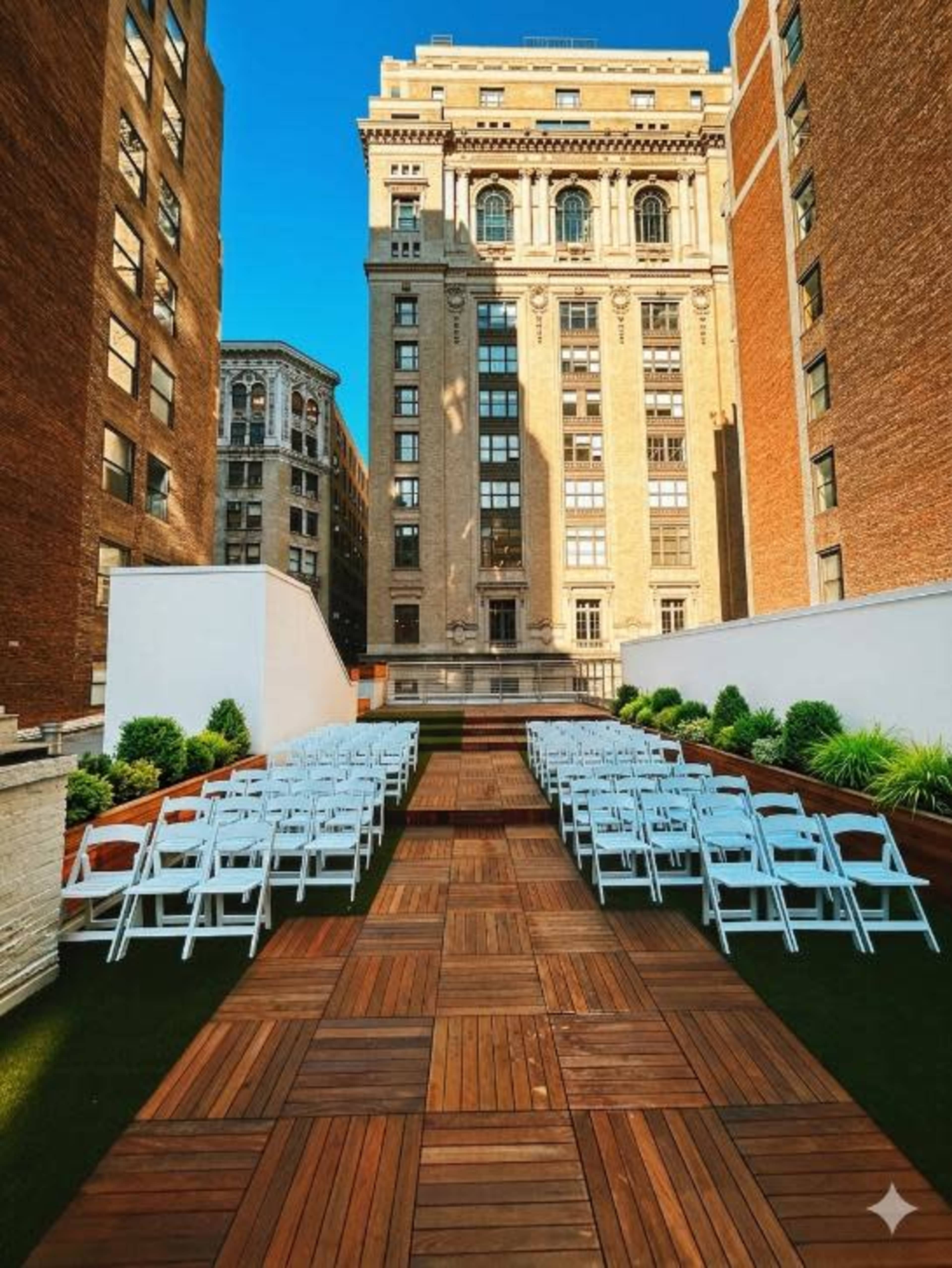 The image shows a rooftop venue with rows of white chairs arranged on a wooden pathway, leading toward a tall building in the background under a clear blue sky.