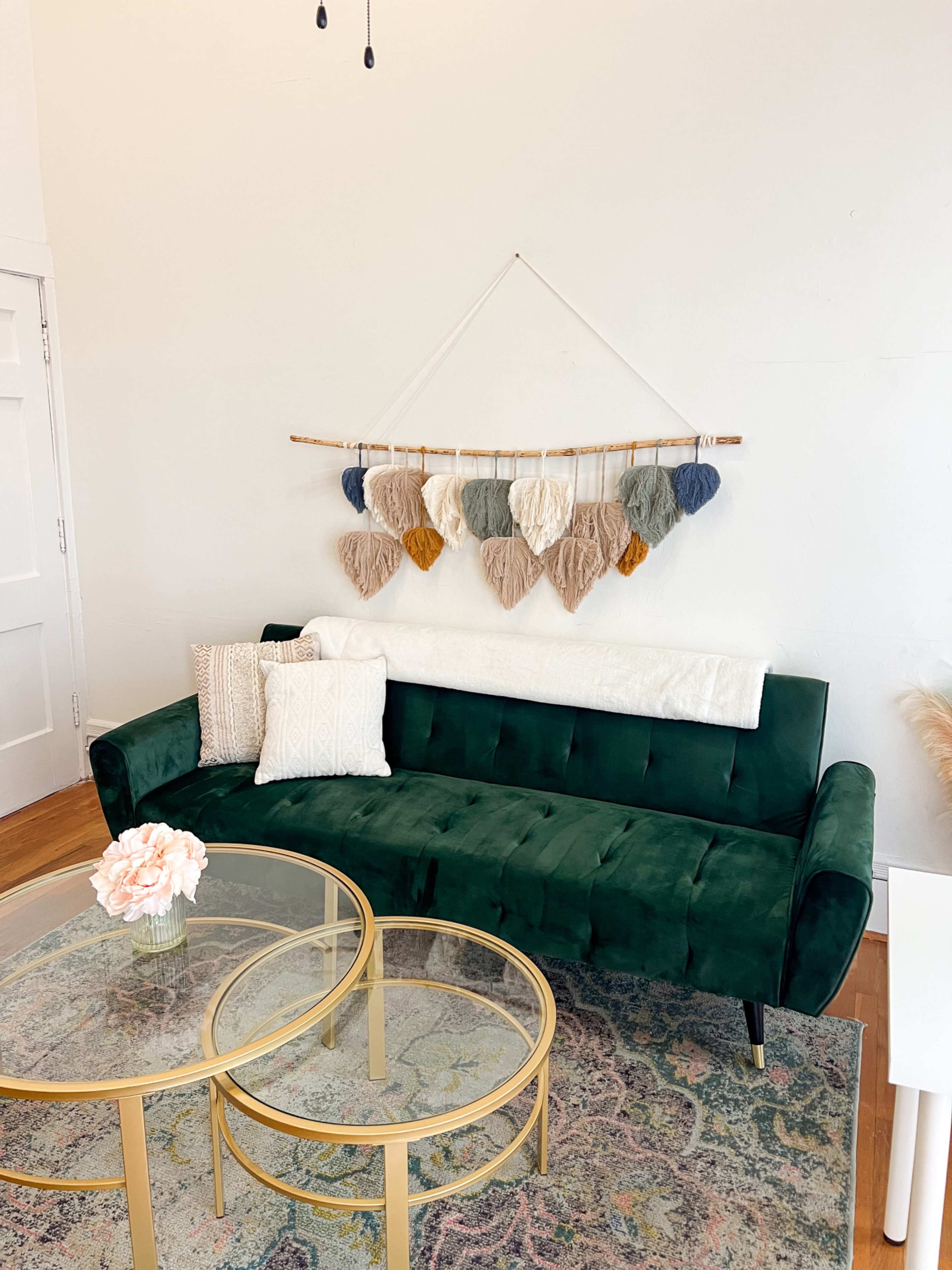 A green velvet sofa is positioned against a white wall, adorned with a hanging wall decoration made of yarn in various colors, with a glass coffee table in front.
