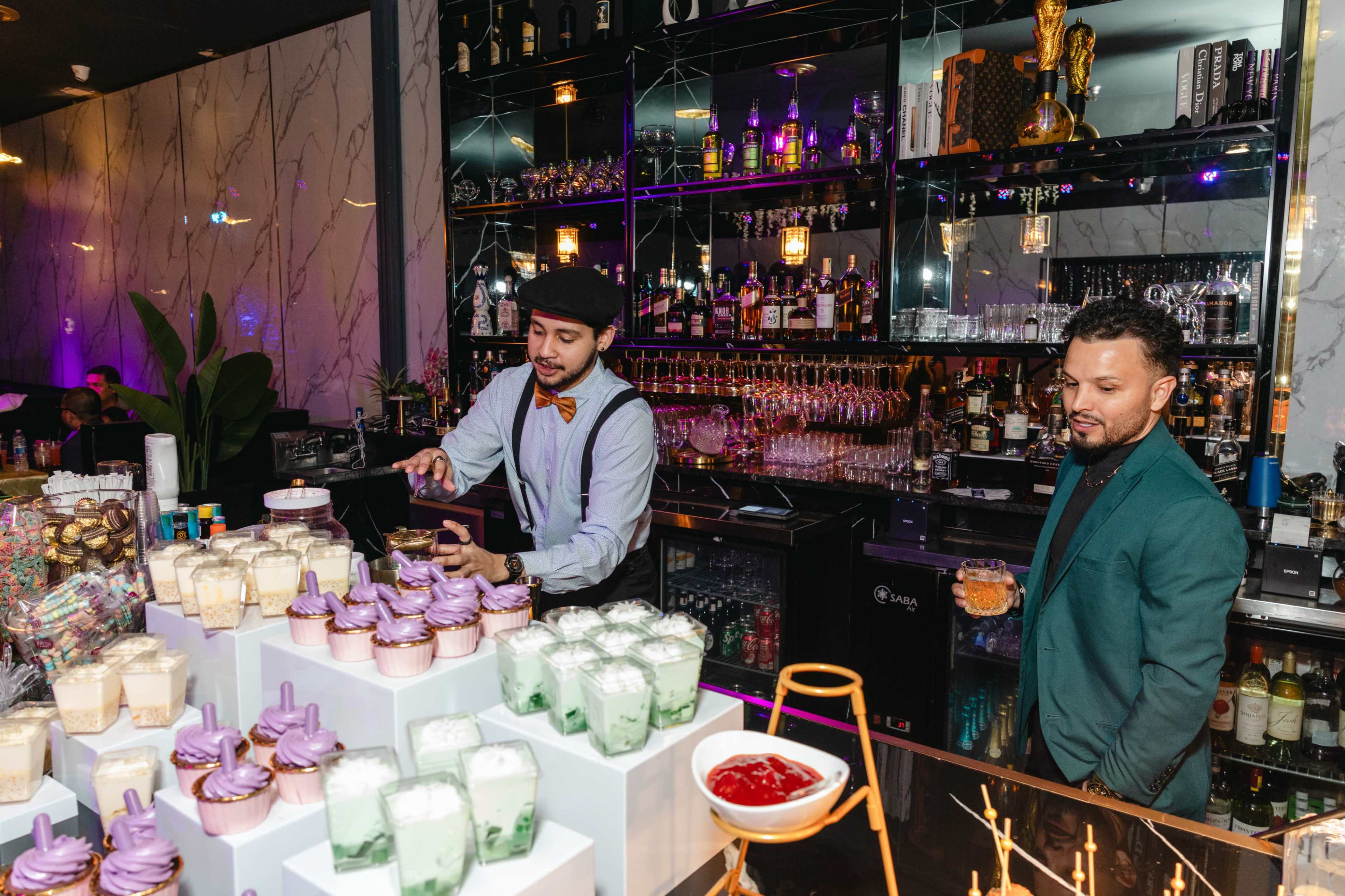A bartender prepares drinks at a stylish bar while a man in a green jacket observes the scene.