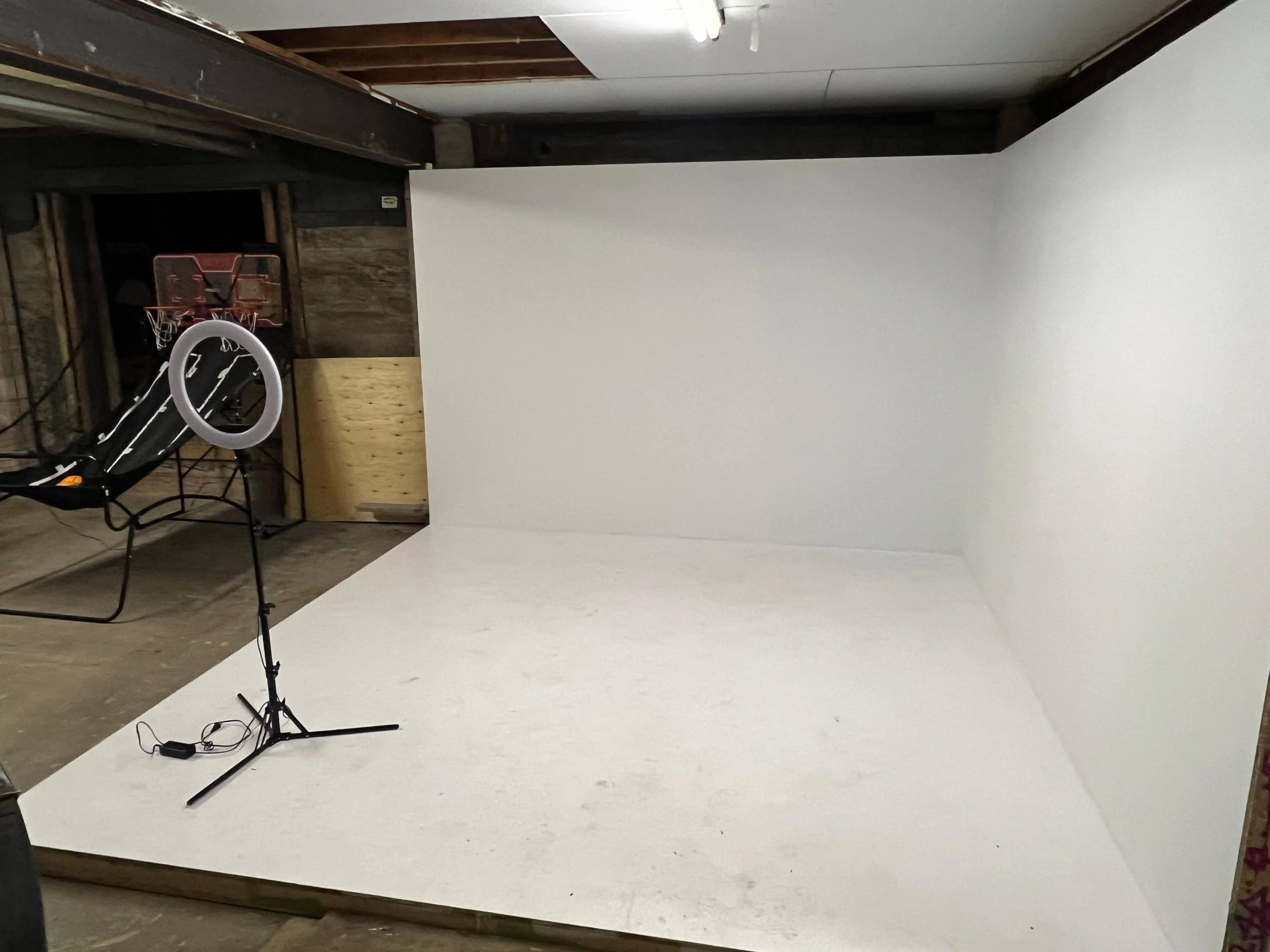 A minimalistic photo studio setup with a white backdrop, a ring light on a stand, and a basketball hoop in the background.