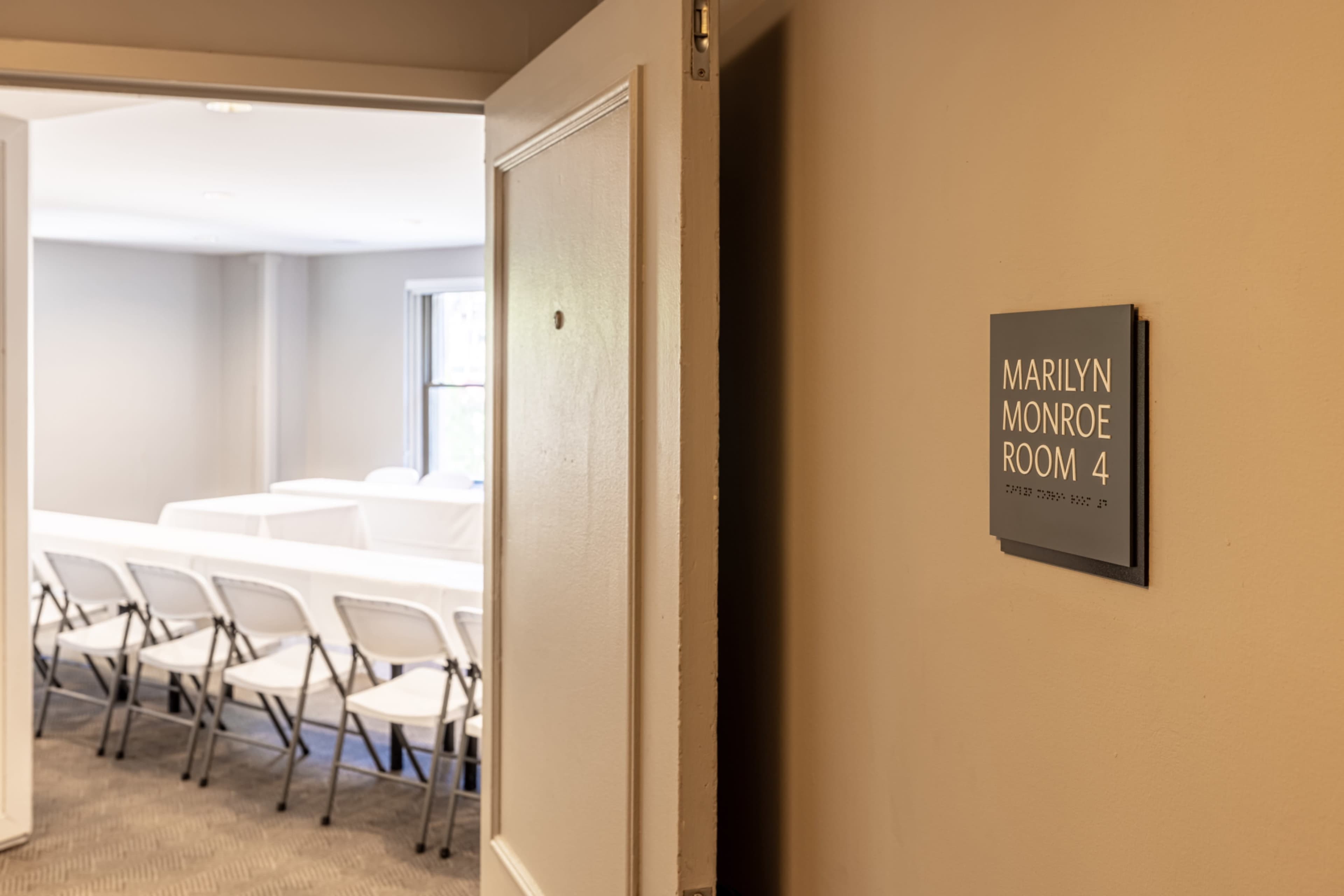 The image shows a sign labeled "Marilyn Monroe Room 4" on a wall next to an open door leading into a meeting space with chairs arranged in rows.