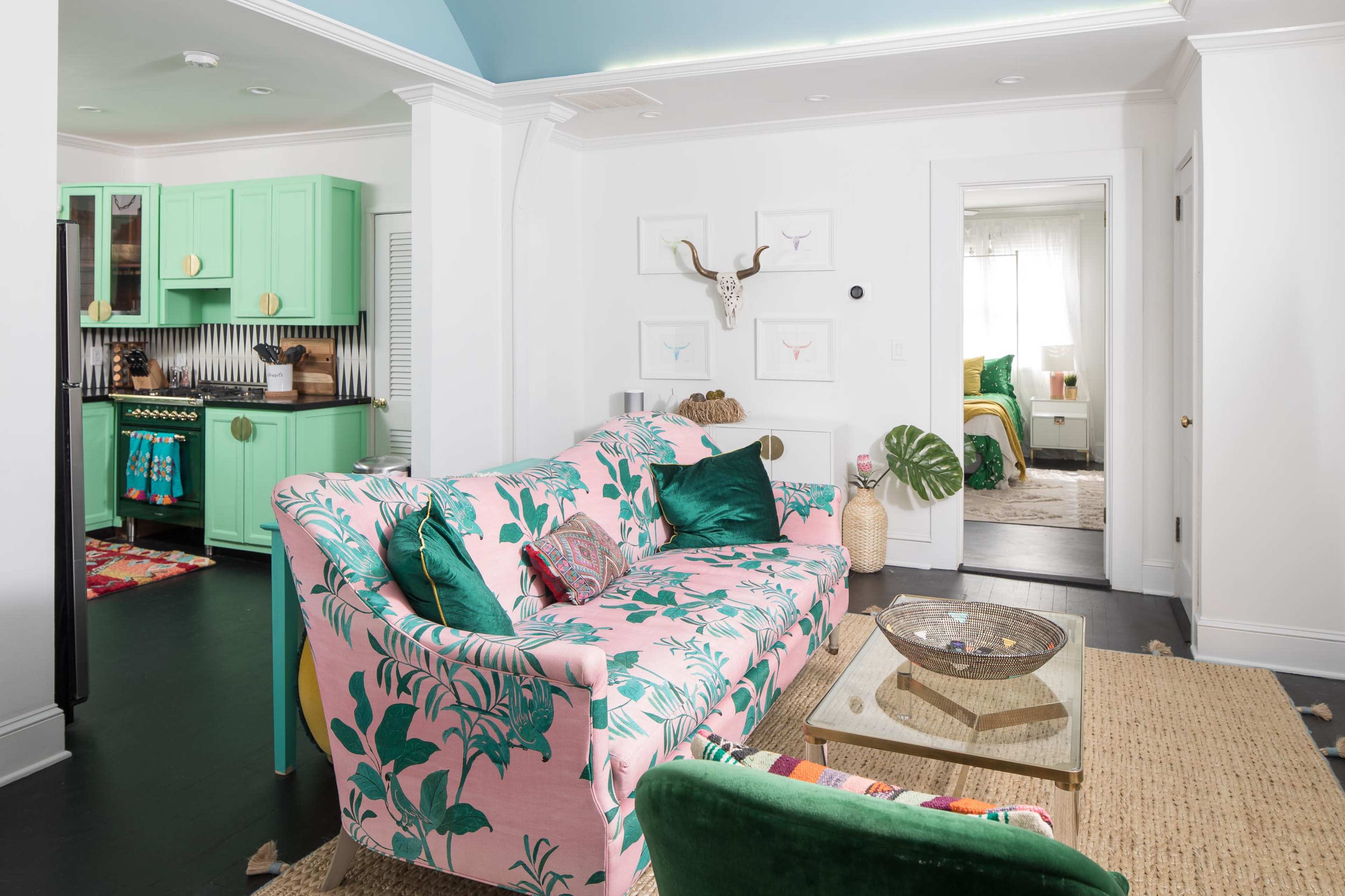 A brightly colored living room features a pink floral-patterned sofa, a glass coffee table, and a view of a green kitchen in the background.