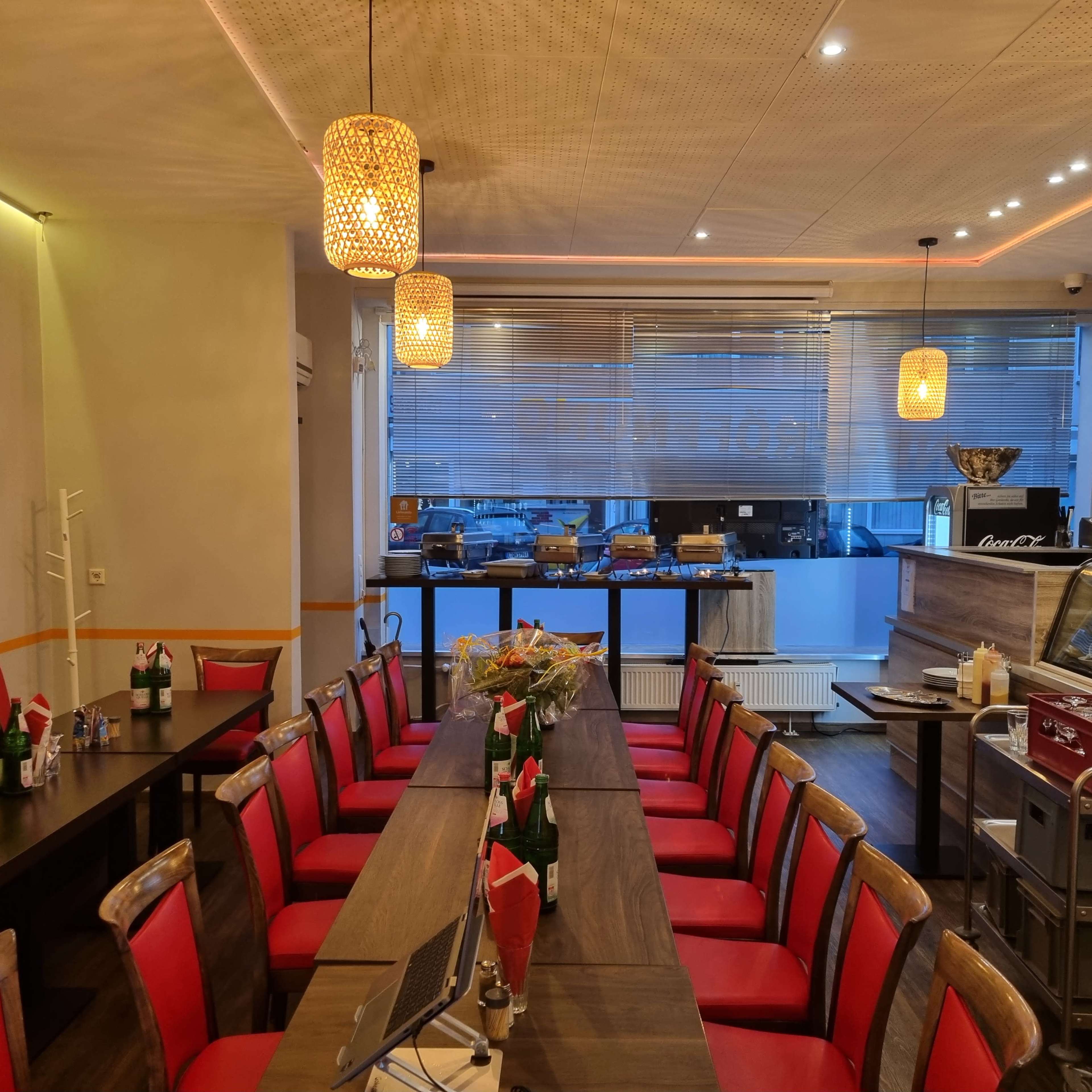 A long wooden table with red chairs is set up in a restaurant, illuminated by hanging pendant lights, with a buffet area visible in the background.